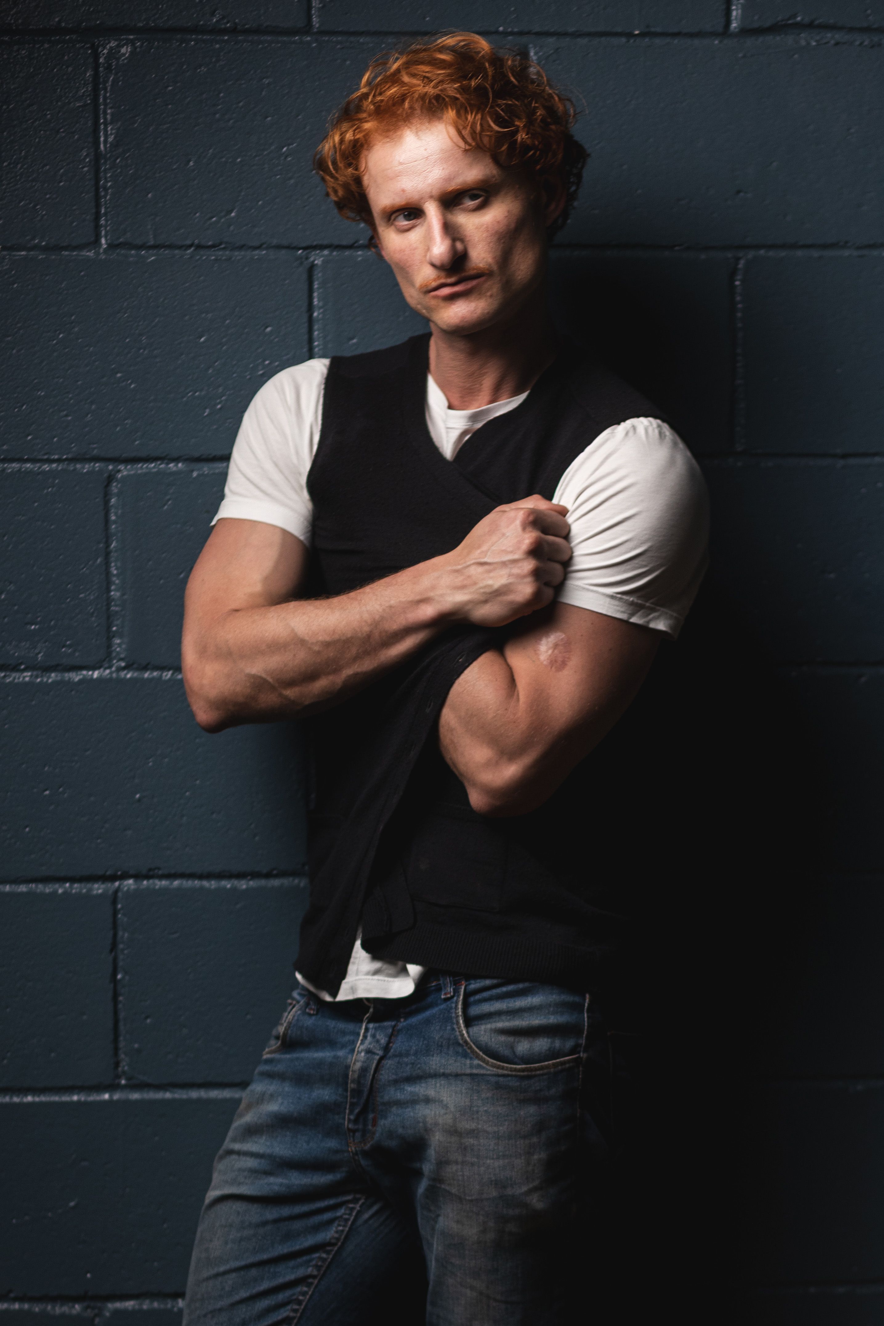 Portrait of a man with red hair and casual attire, leaning against a wall.