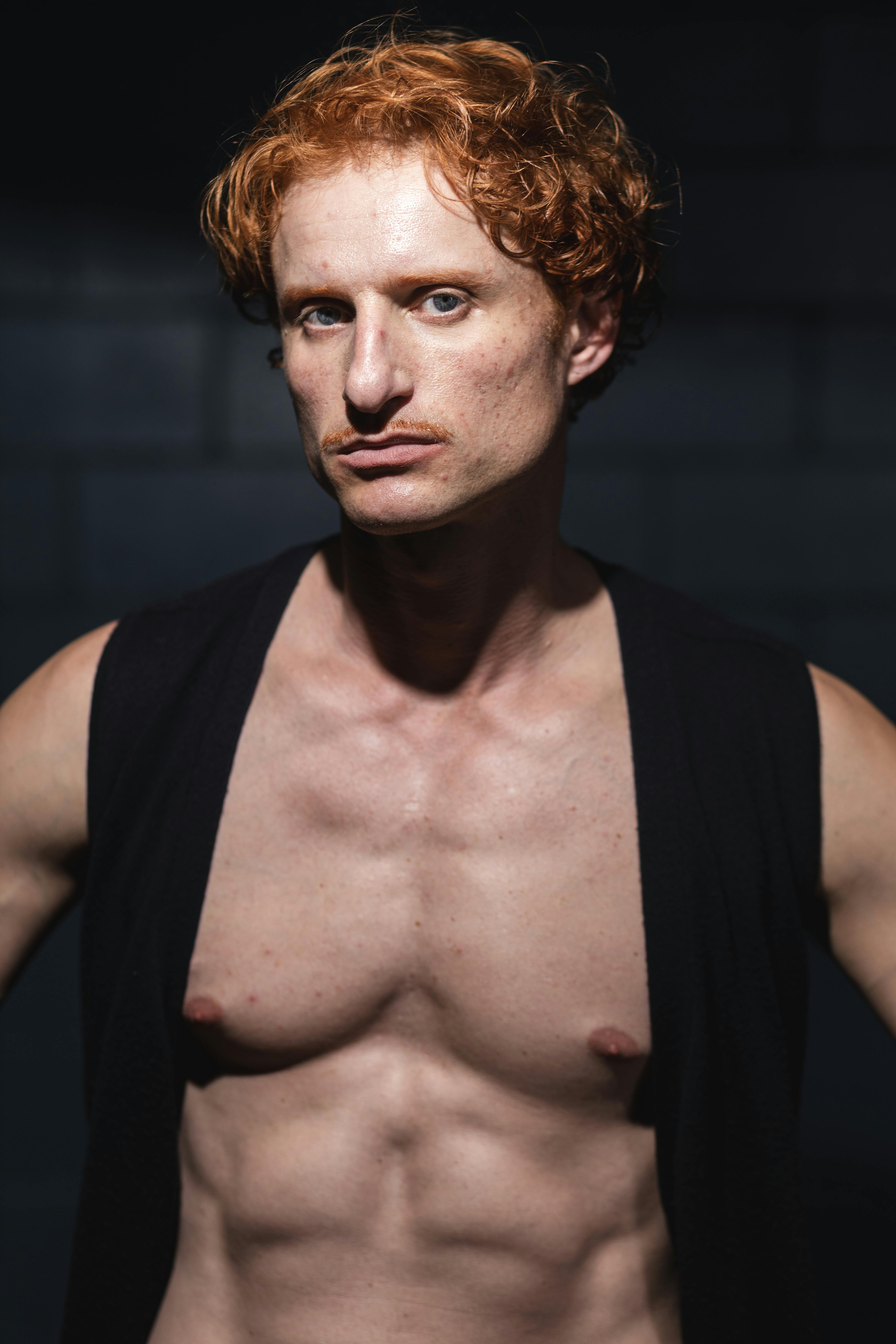 Muscular man with red hair and serious expression posing in a dark studio setting.
