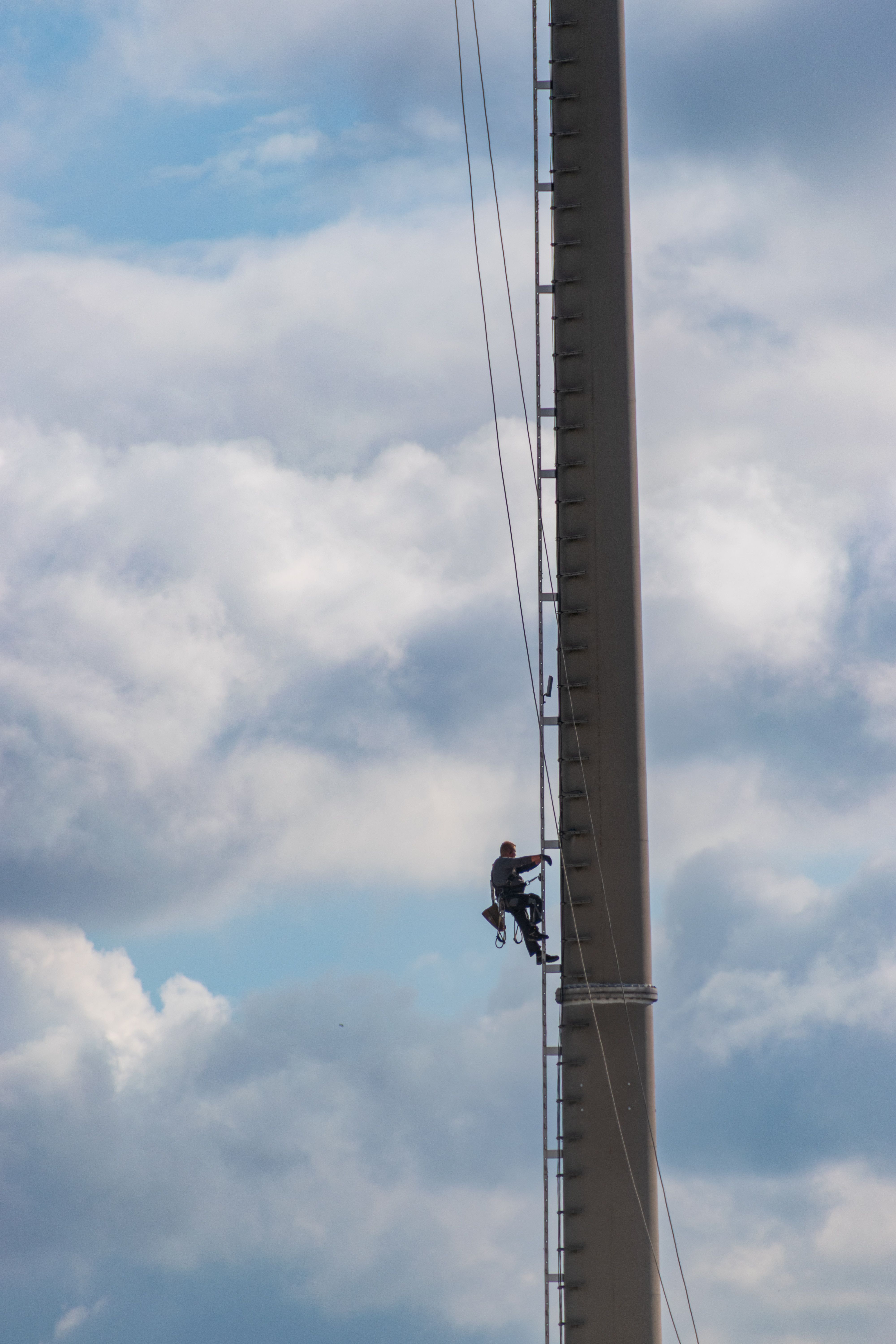 A worker climbs a telecommunication tower against a backdrop of cloudy skies, showcasing industrial safety.