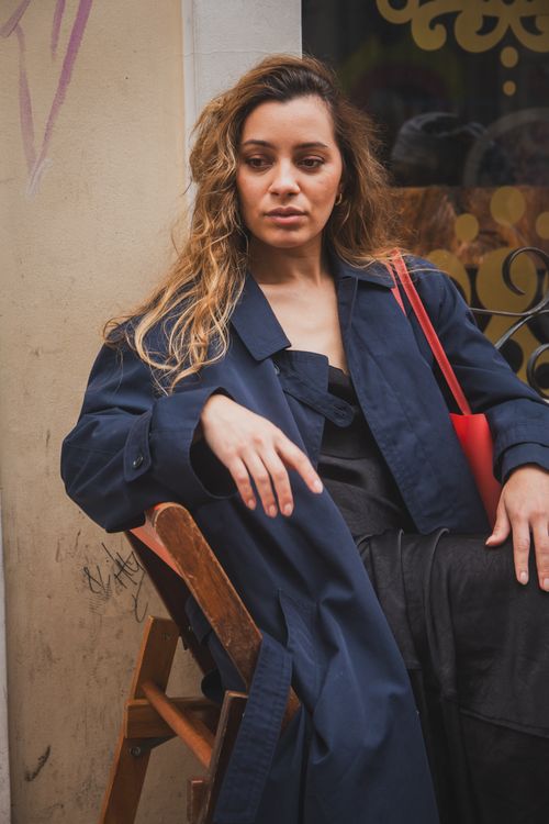 Free A fashionable woman in a blue coat and red bag poses confidently on a wooden chair outdoors. Stock Photo