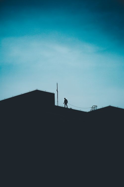 Free A solitary silhouette stands atop a building against a blue sky, evoking themes of courage and solitude. Stock Photo