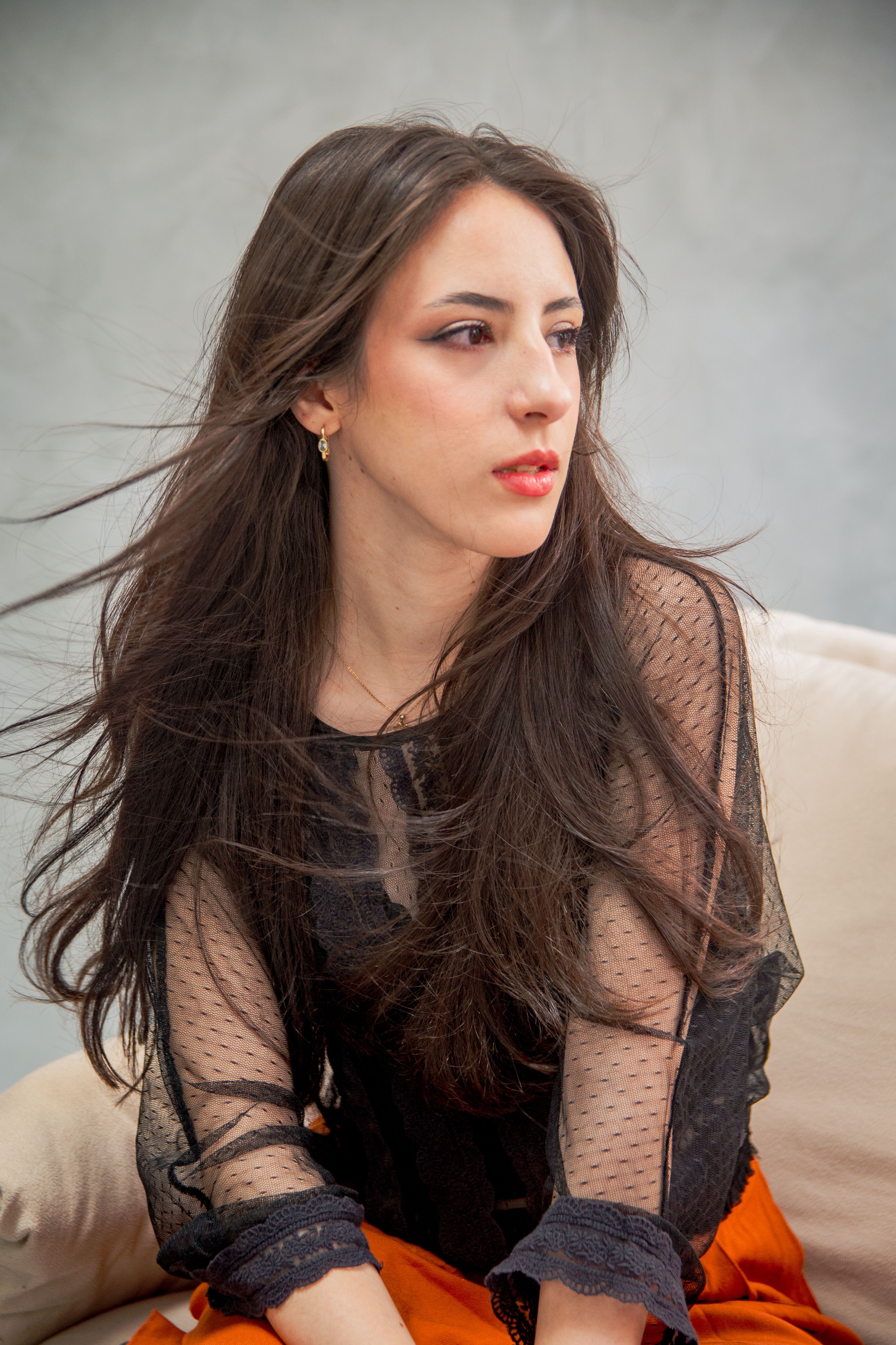 Portrait of a woman with long flowing hair in a sheer black top and orange skirt, gazing thoughtfully.
