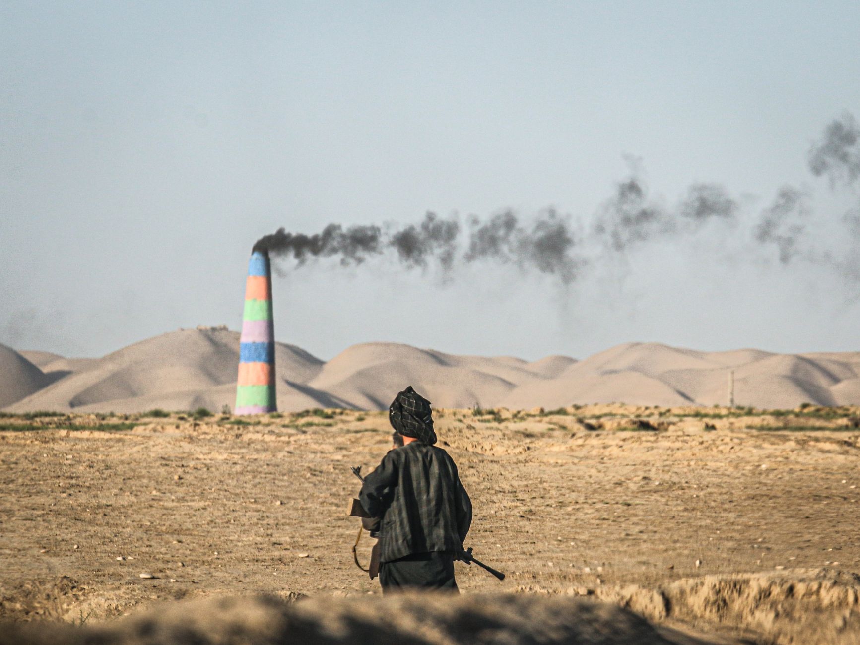 A man stands watching smoke billow from a chimney in the Afghanistan desert landscape.
