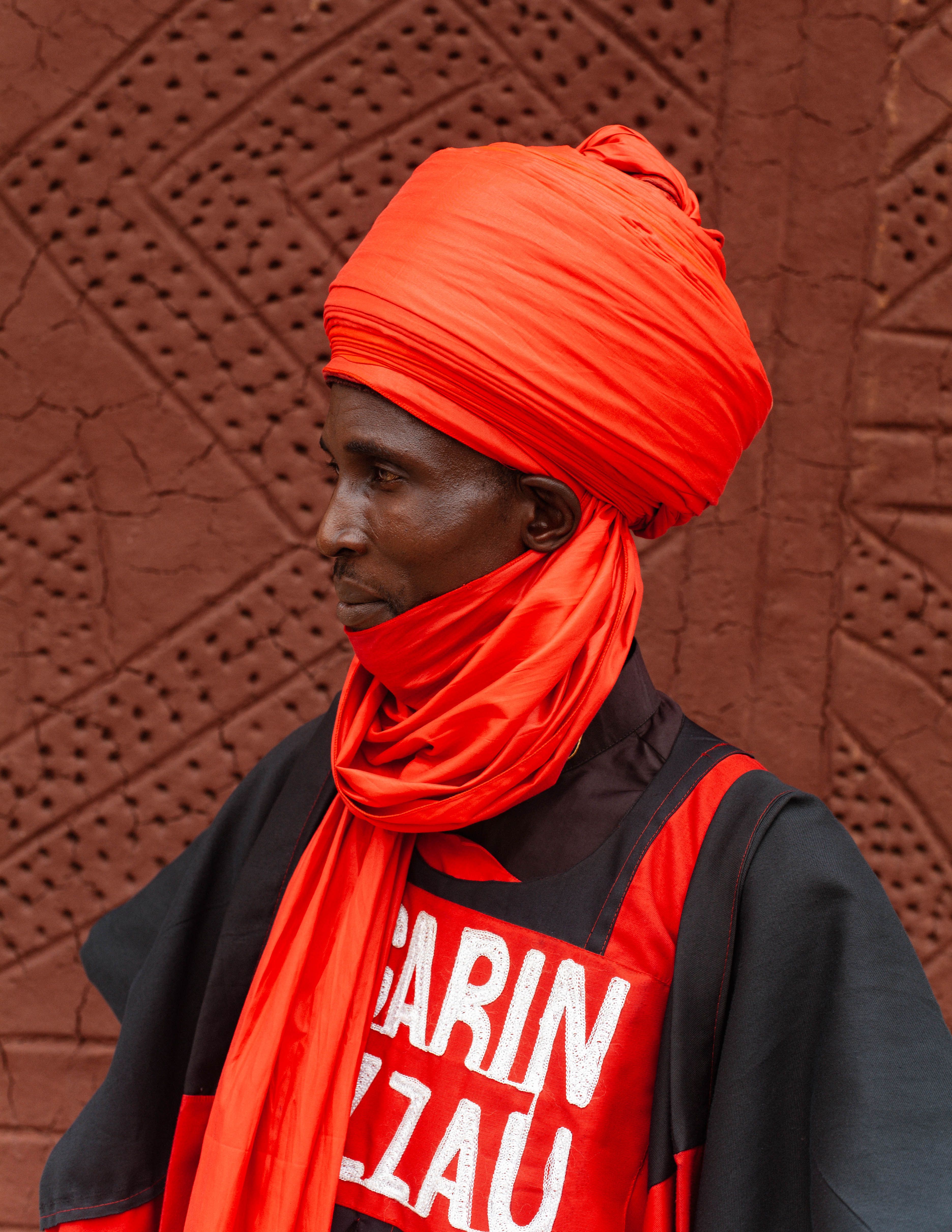 A man in vibrant red traditional attire against an intricate patterned wall.