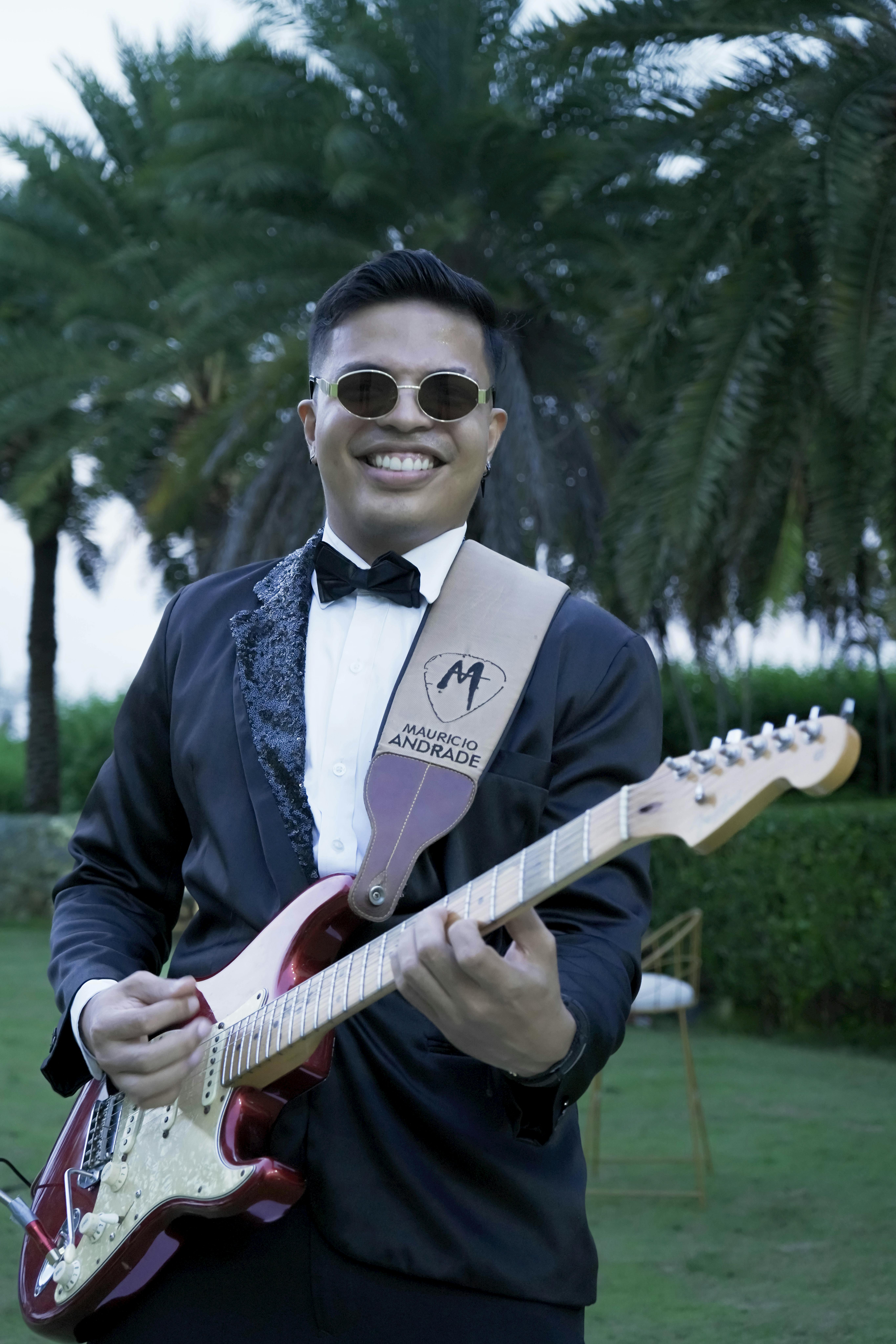Smiling man playing electric guitar outdoors in a tuxedo with palm trees in the background.