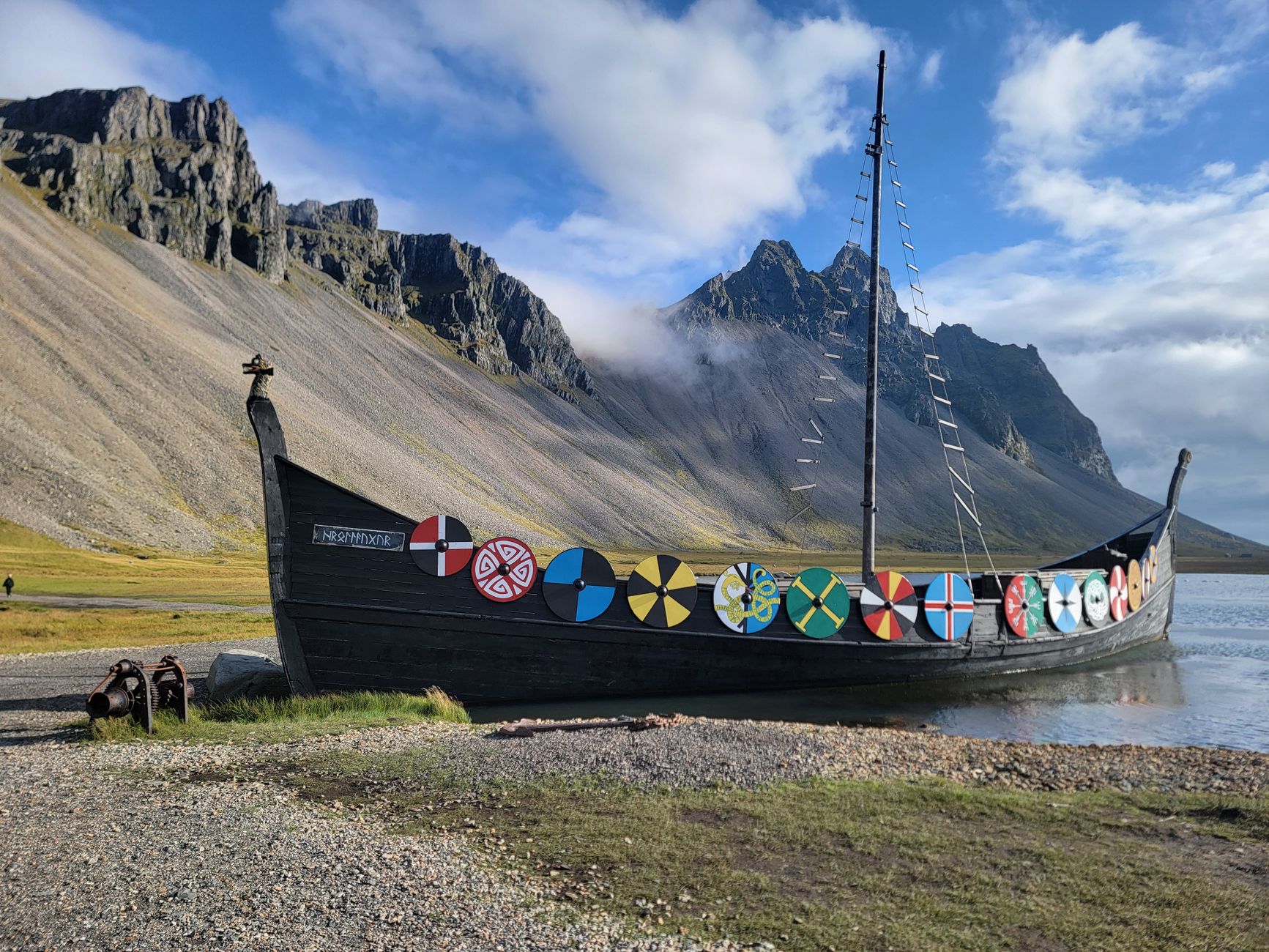 Traditional Viking ship with colorful shields docked by a rugged mountain coastline under a bright sky.