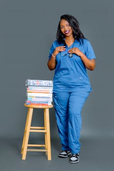 Happy female nurse in blue scrubs with stethoscope and textbooks on stool.