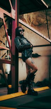 Stylish individual in punk attire with fishnet stockings posing in an industrial setting.