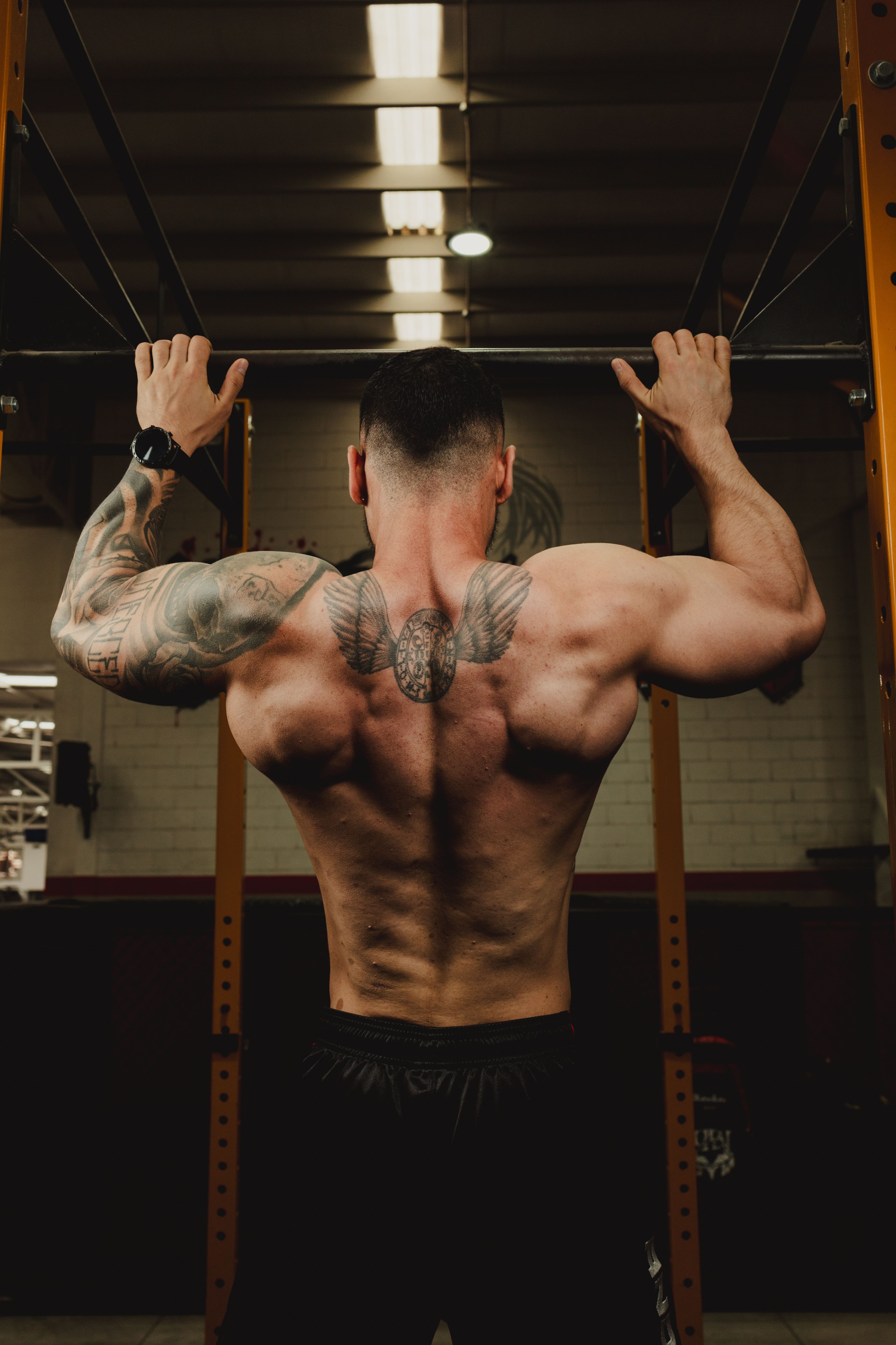 Back view of a man with tattoos doing pull-ups in a gym setting, showing strength and motivation.
