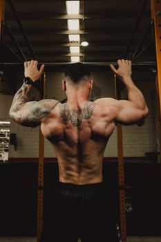 Back view of a man with tattoos doing pull-ups in a gym setting, showing strength and motivation.