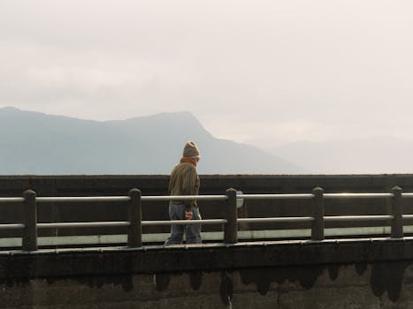 An elderly man in warm clothing walks on a bridge in Scotland during sunset, with mountains in the background.