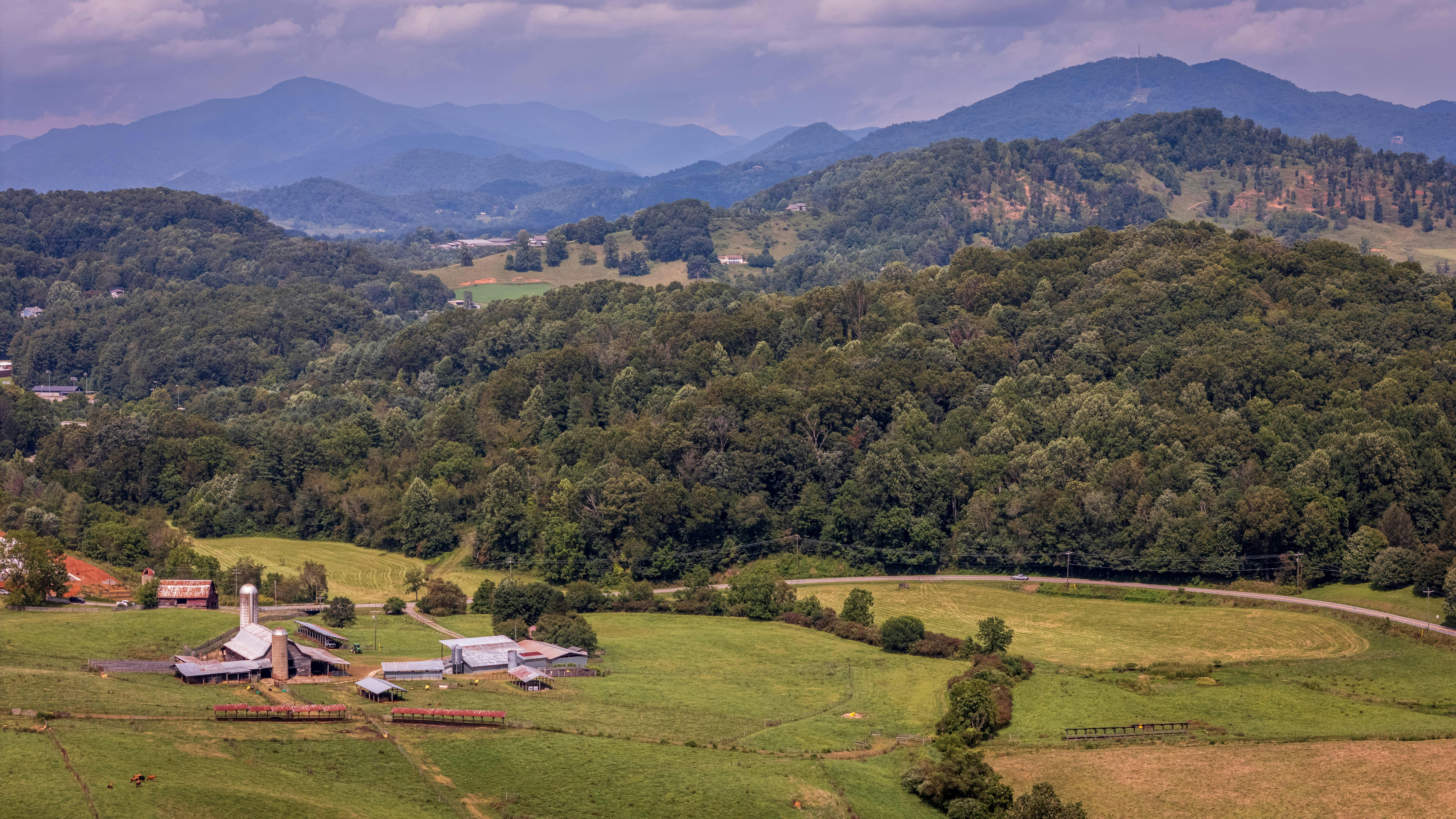Aerial view of a picturesque farm surrounded by forested hills and distant blue mountains.