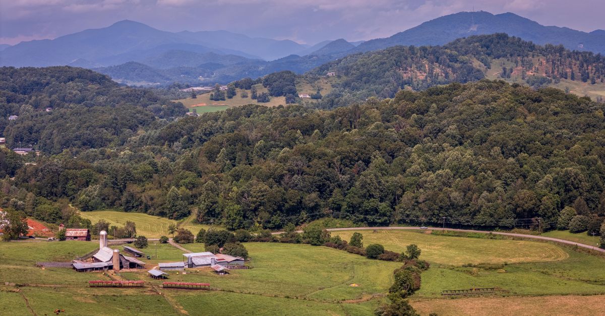 Aerial view of a picturesque farm surrounded by forested hills and distant blue mountains.