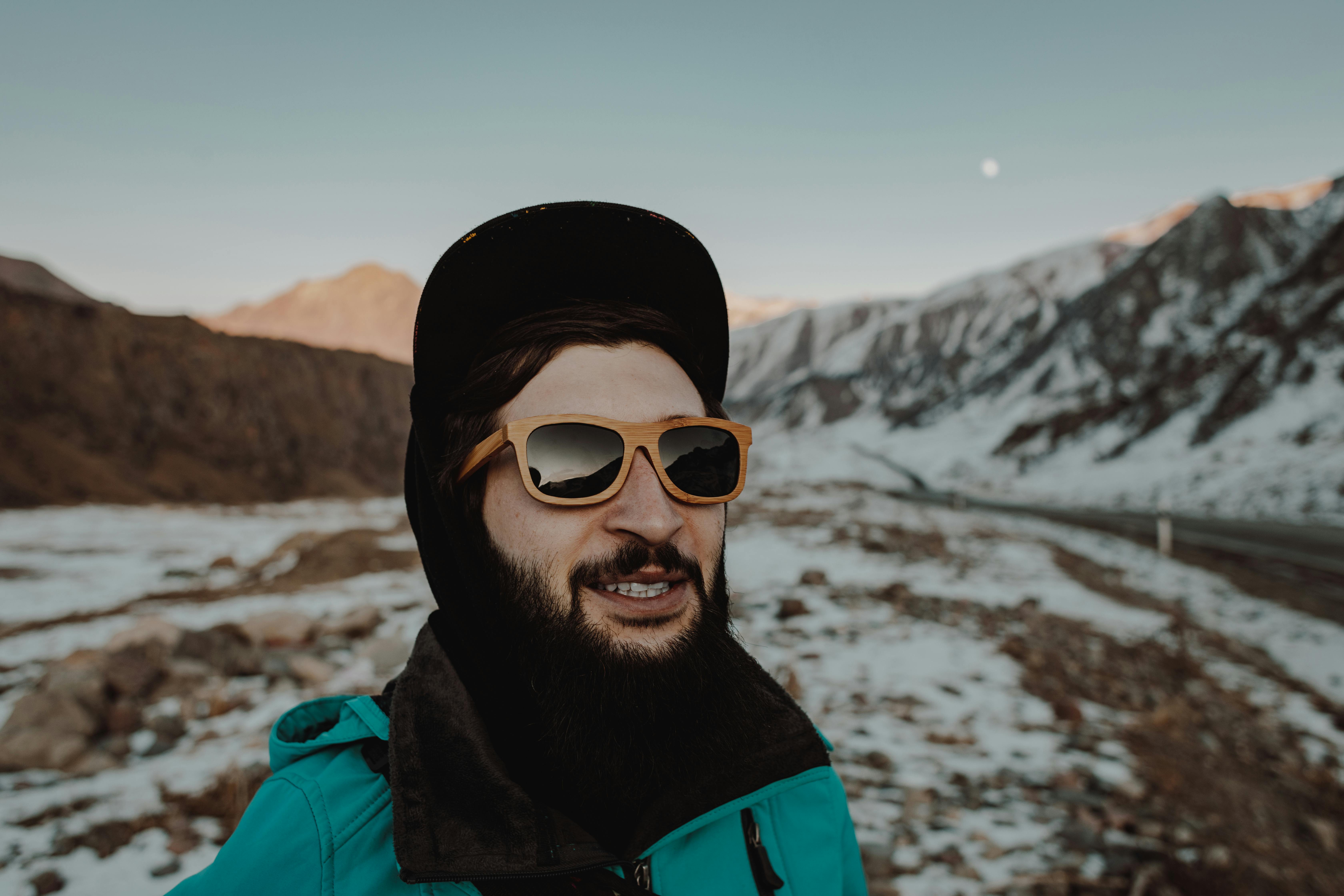 在户外雪山风景中，一位脸上有胡须、戴着太阳镜的男子微笑着