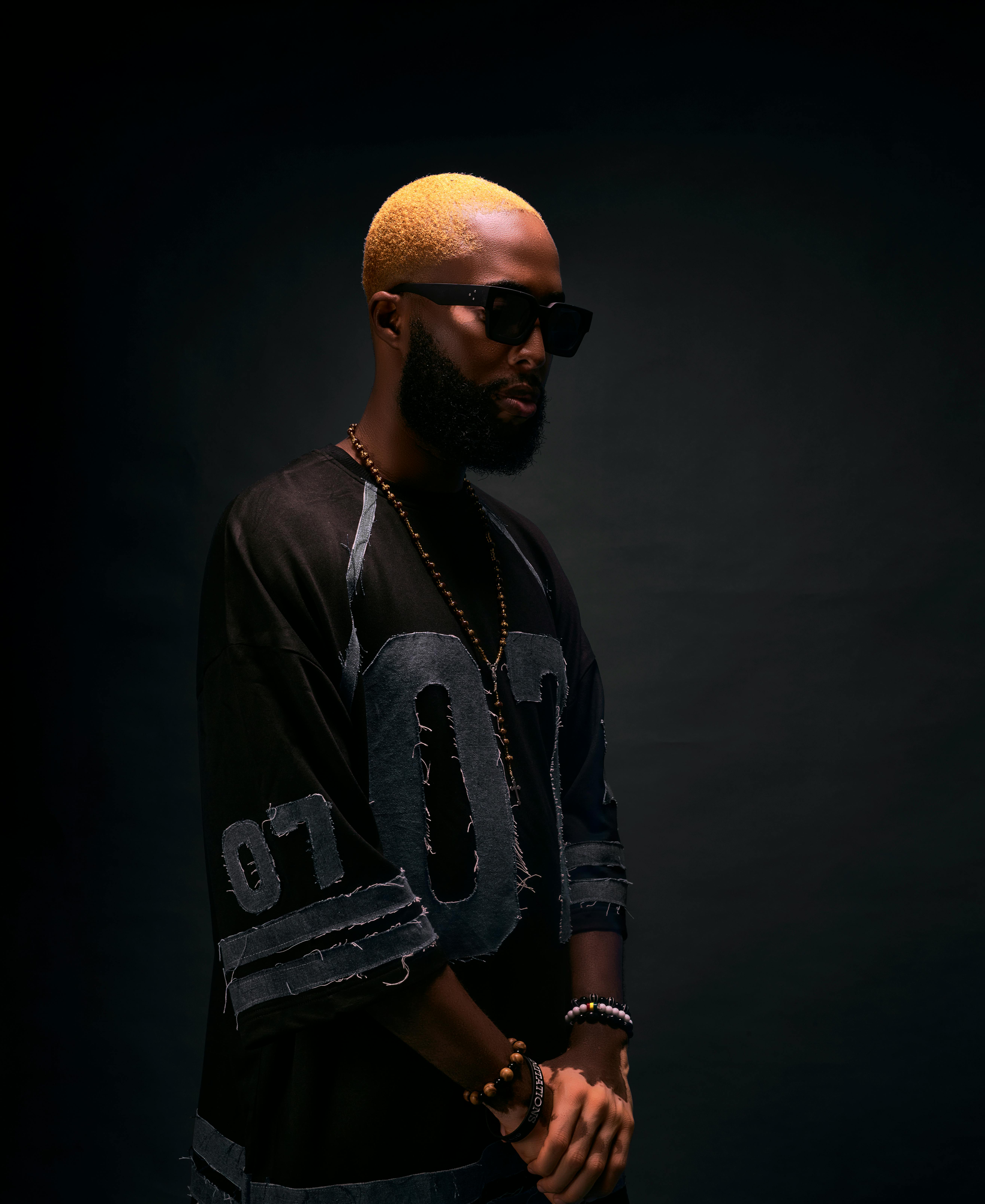 Contemporary portrait of a man with blonde hair, sunglasses, and a beard against a dark backdrop.