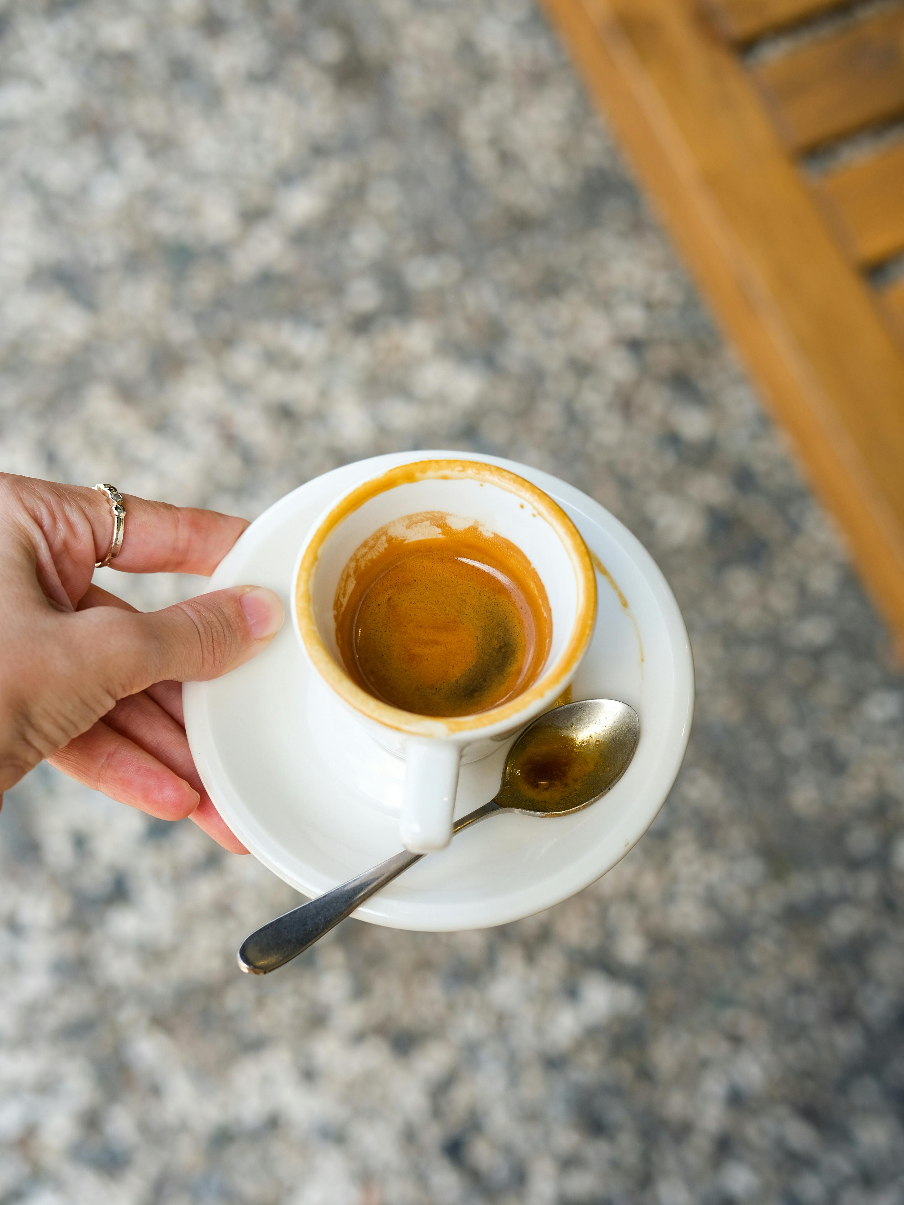 A close-up of a hand holding a white espresso cup on a saucer with a spoon, viewed from above.