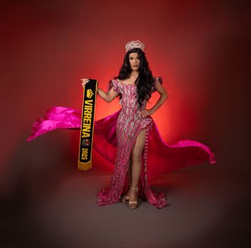 Glamorous pageant winner in shimmering gown and crown holding a banner.