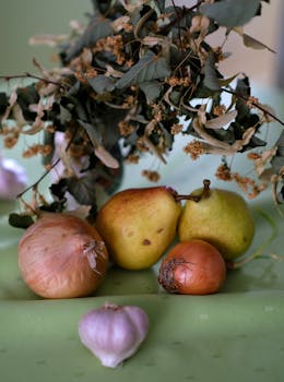 A rustic still life featuring pears, onions, garlic, and dried leaves on a green surface.