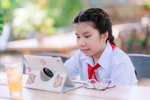 Young girl in school uniform studying on tablet outside, with drink and glasses beside her.