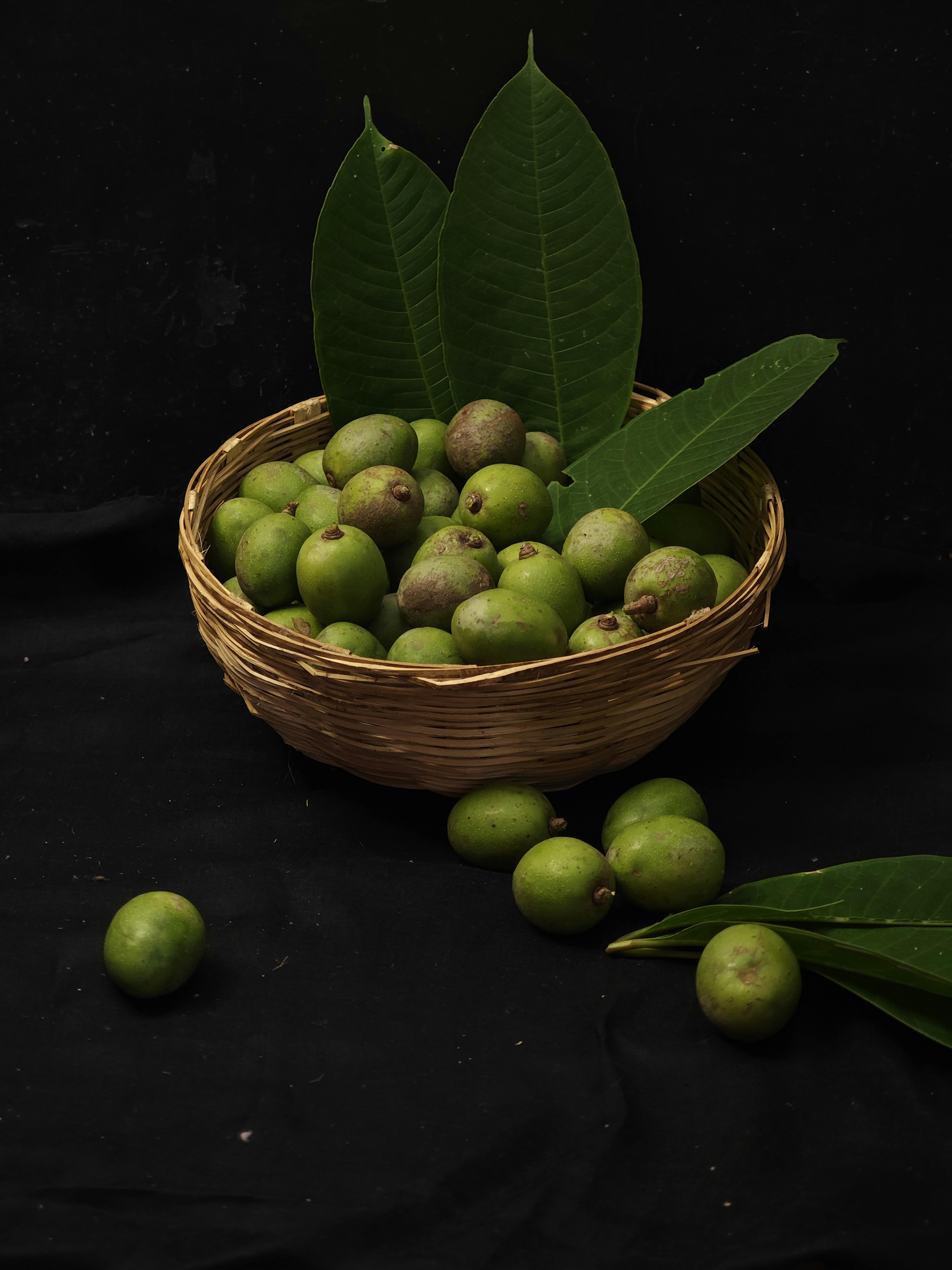 A wicker basket filled with fresh green mangoes and leaves on a black background, offering a rich and earthy visual.