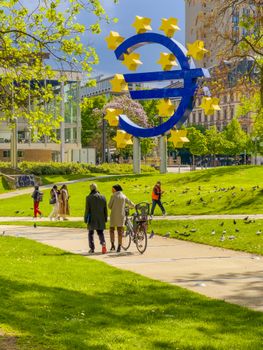 People walking near the iconic Euro sculpture in a sunny Frankfurt park.
