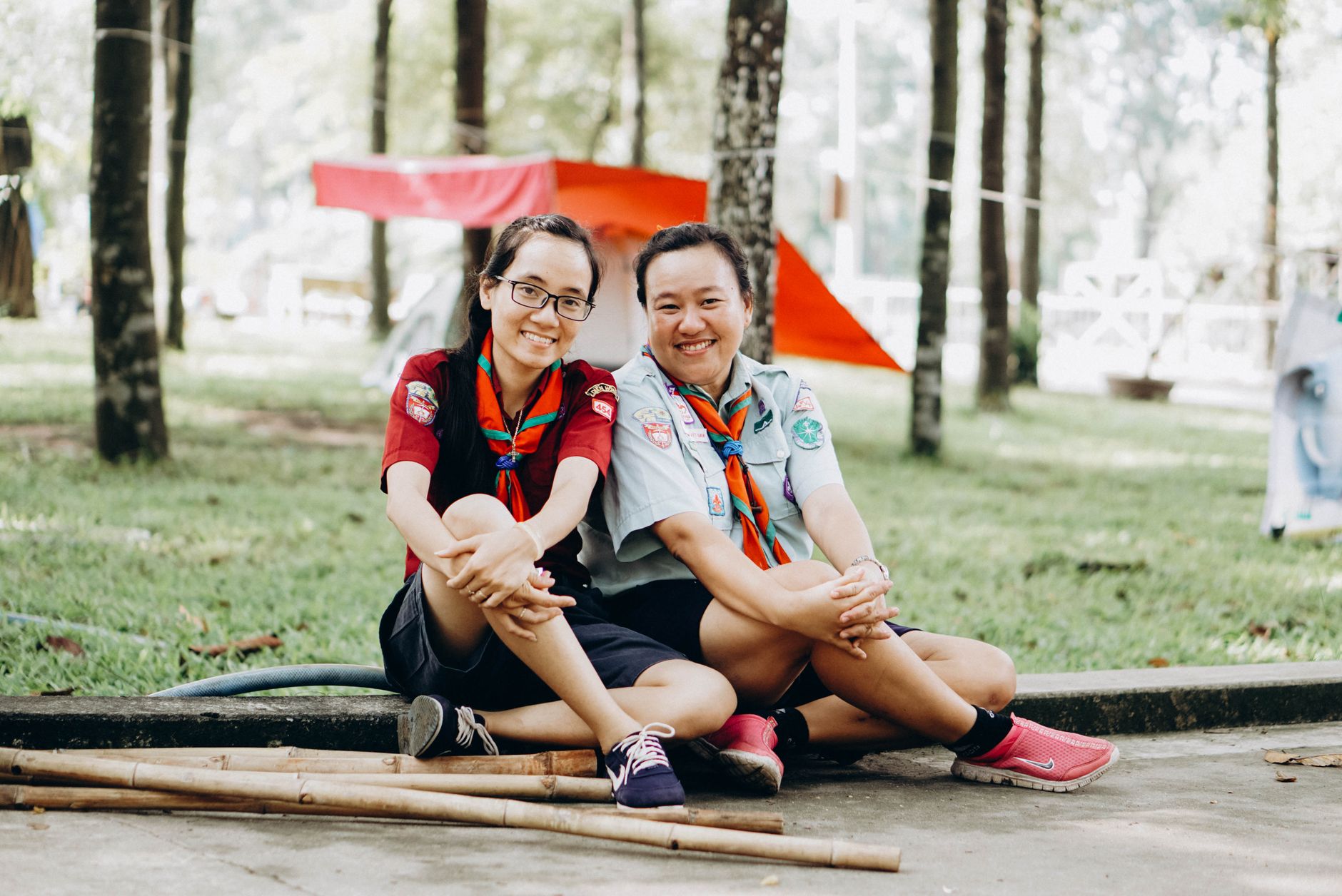 Two cheerful scouts sitting on grass in a campsite, surrounded by trees and tents.
