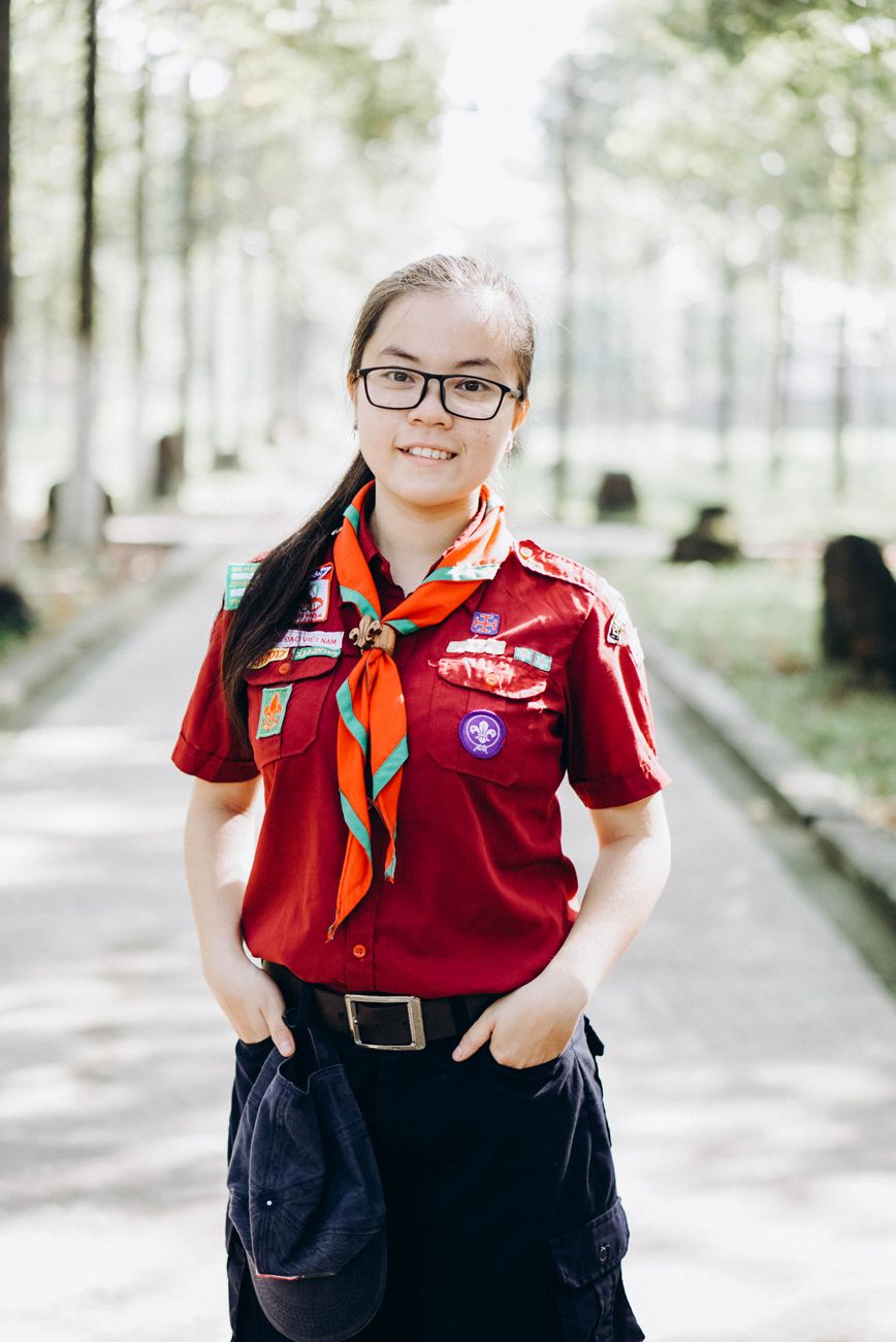 Escoteiro sorridente em uniforme vermelho com distintivos e cachecol em pé em um caminho ensolarado.