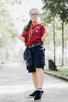 Smiling young scout in a red uniform standing outdoors on a sunny day with trees in the background.