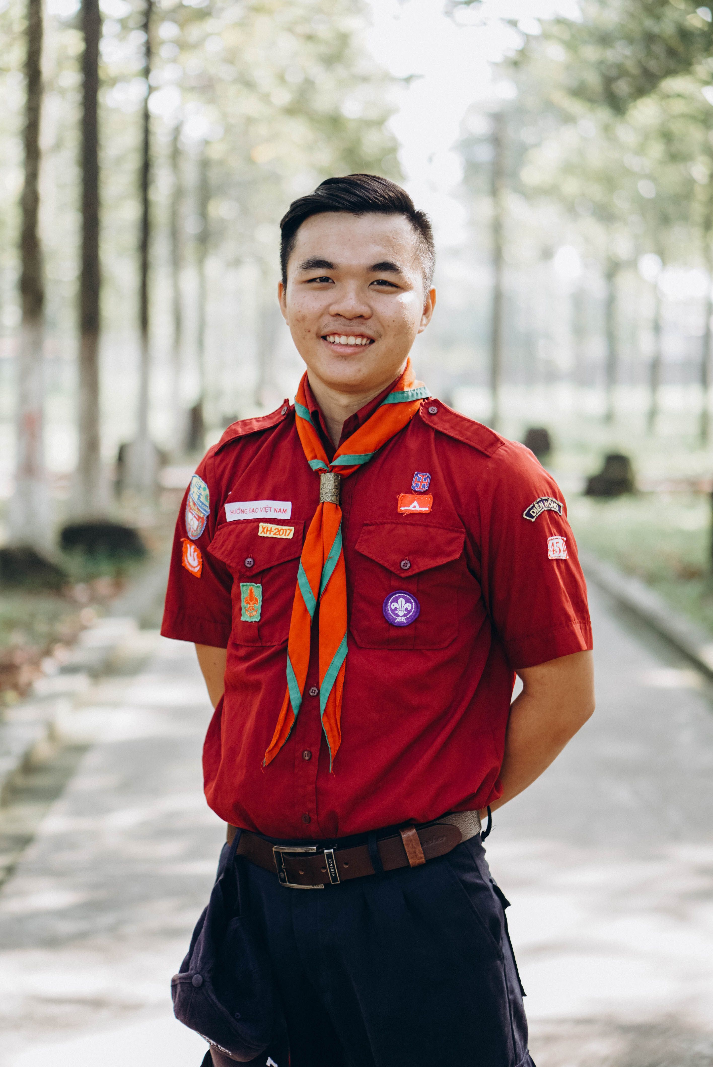 Smiling young adult scout wearing a uniform and scarf, standing outdoors on a sunny day.