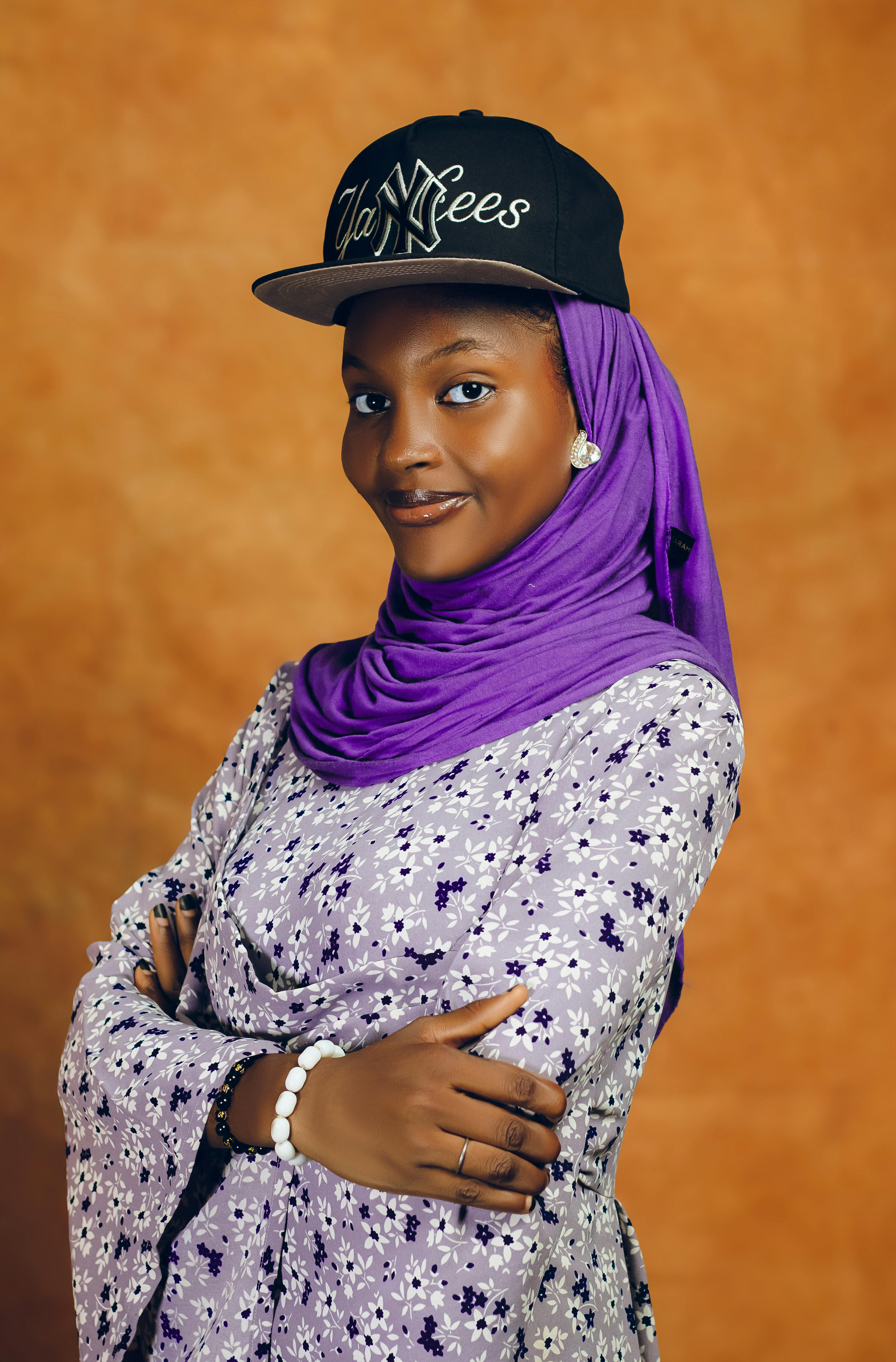 Portrait of a smiling woman wearing a purple hijab and black cap, arms crossed.