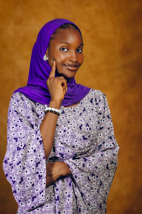 Free Portrait of a smiling woman in purple hijab and floral attire with warm background. Stock Photo