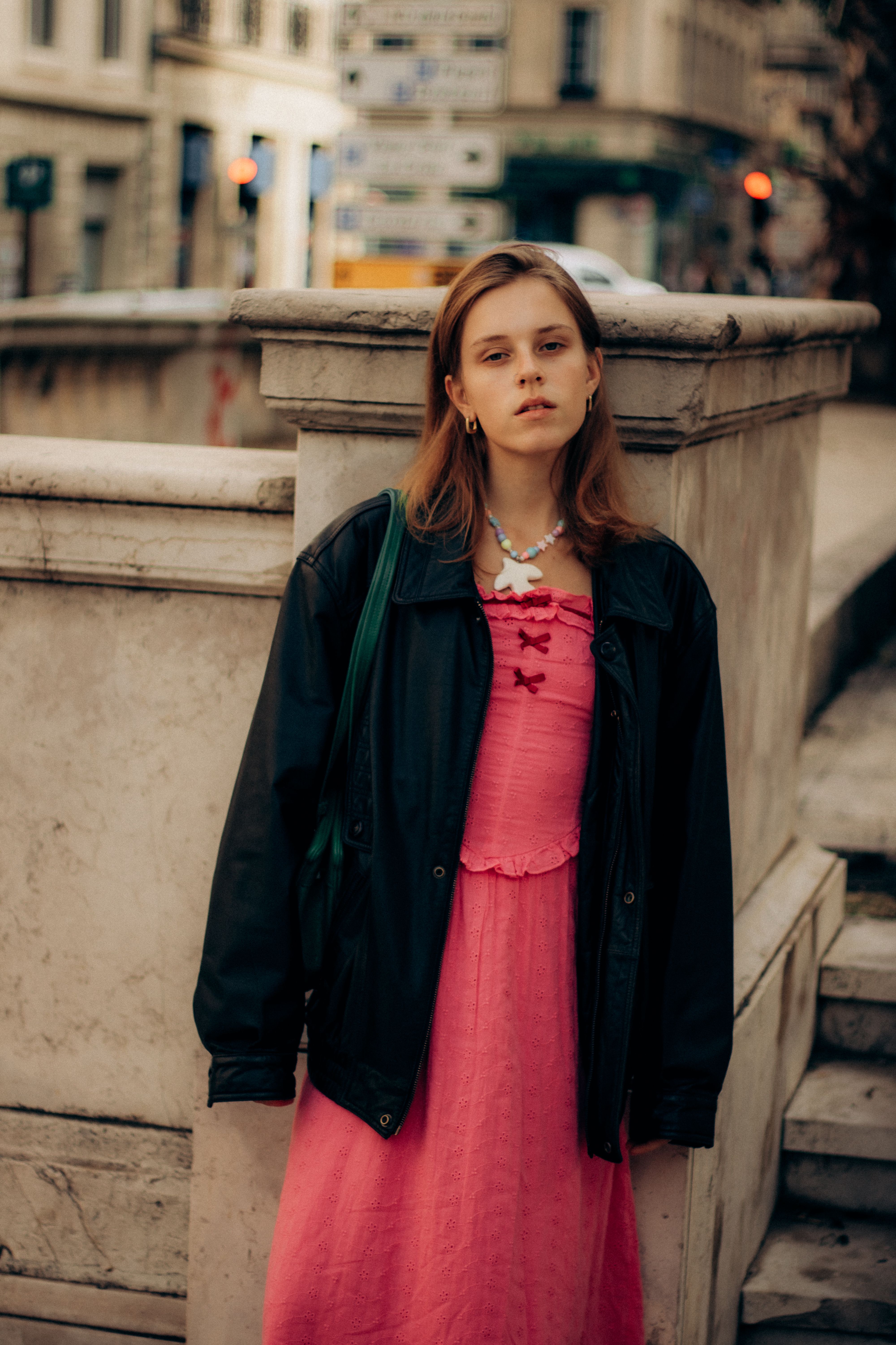 Young woman in a vibrant pink dress and leather jacket leaning on a stone railing in Marseille.
