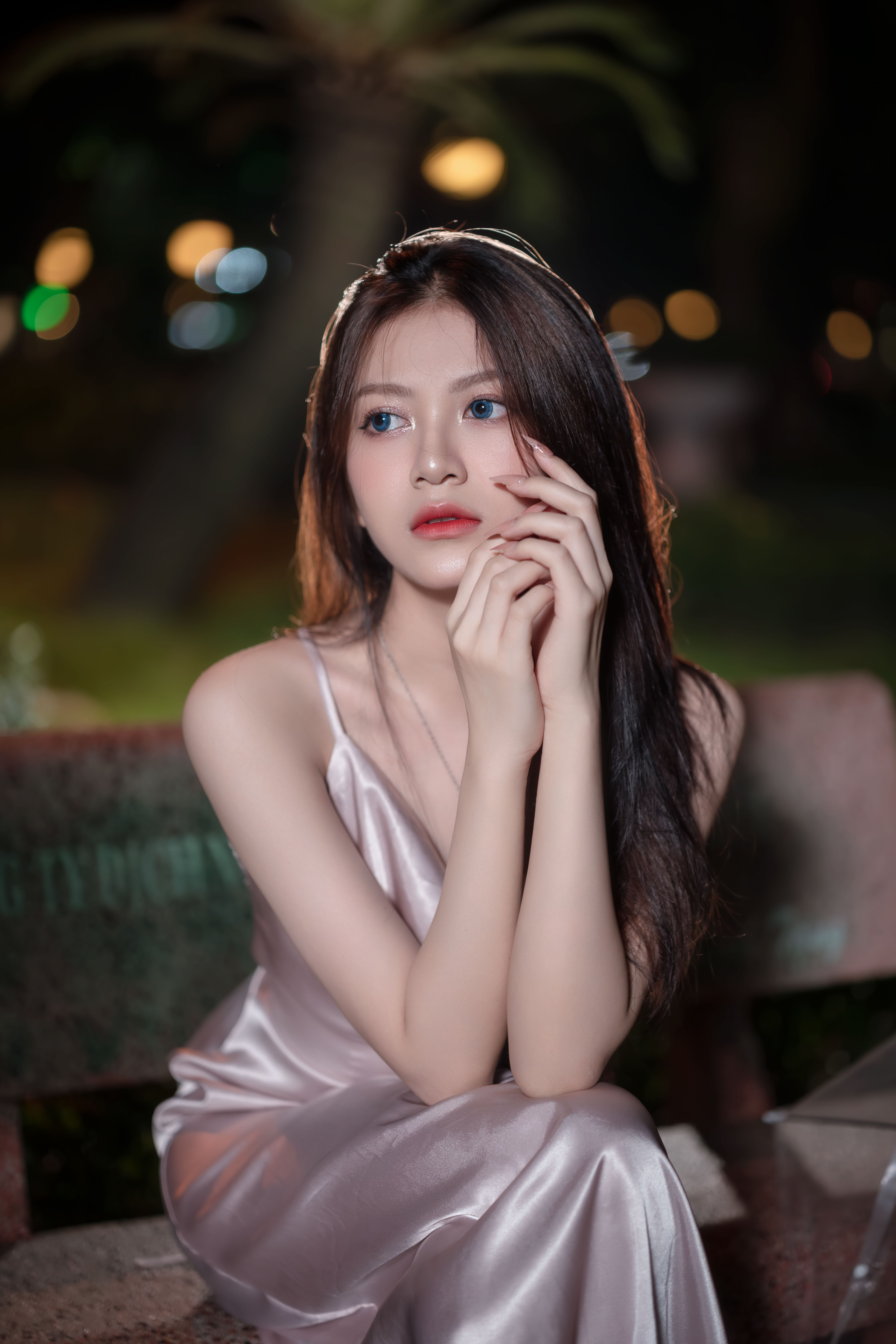 Elegant woman in a satin dress posing thoughtfully on a park bench at night.
