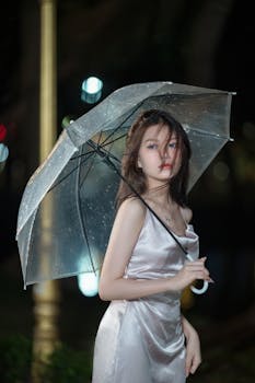 A woman stands elegantly under a transparent umbrella during a rainy night.