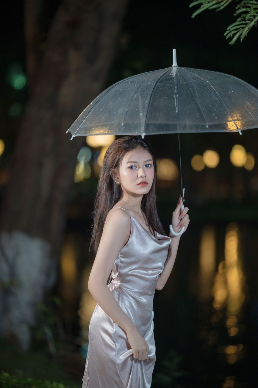 A woman in a satin dress holding a transparent umbrella against a nighttime backdrop.