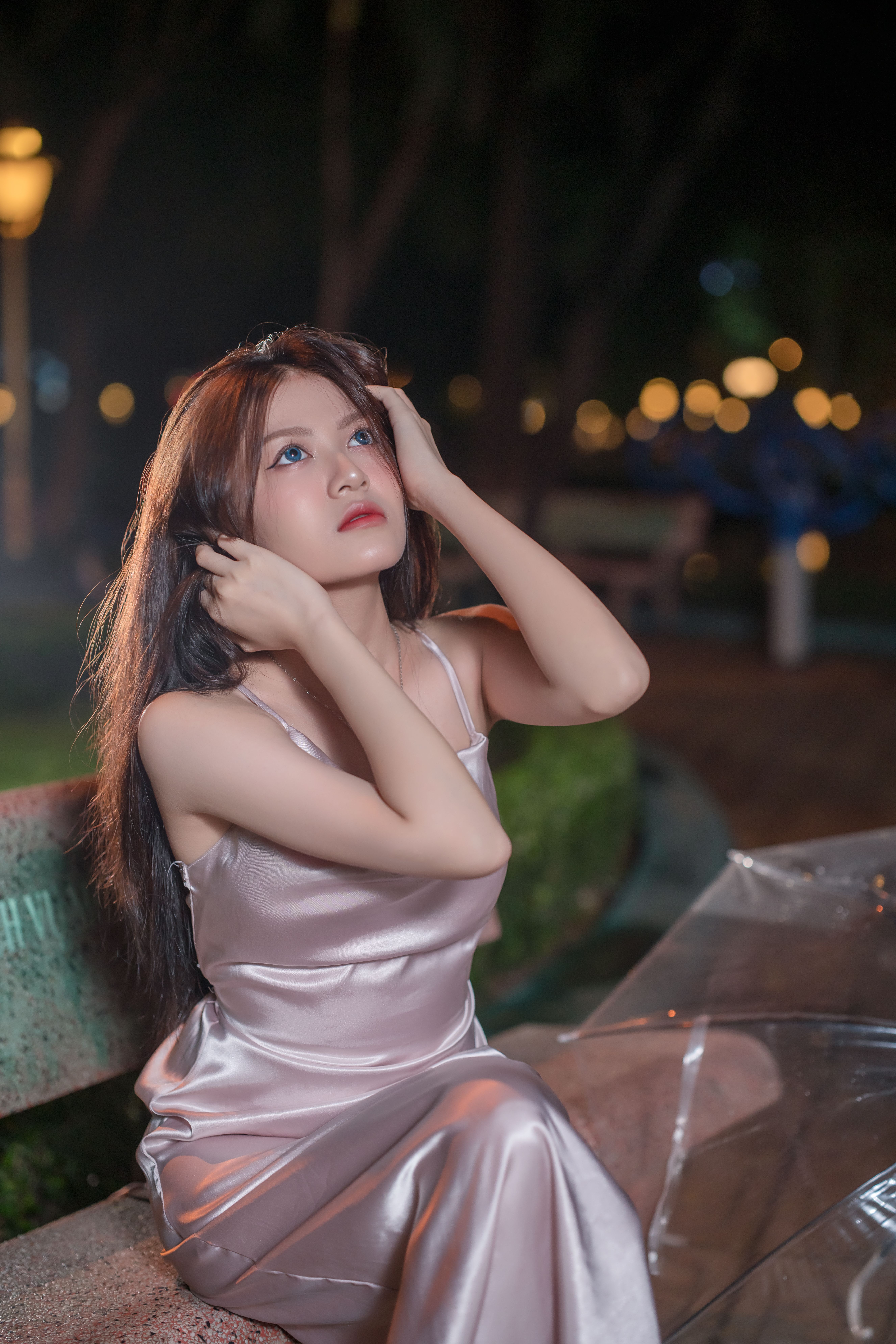 Woman in a satin dress poses on a park bench at night, surrounded by glowing bokeh lights.