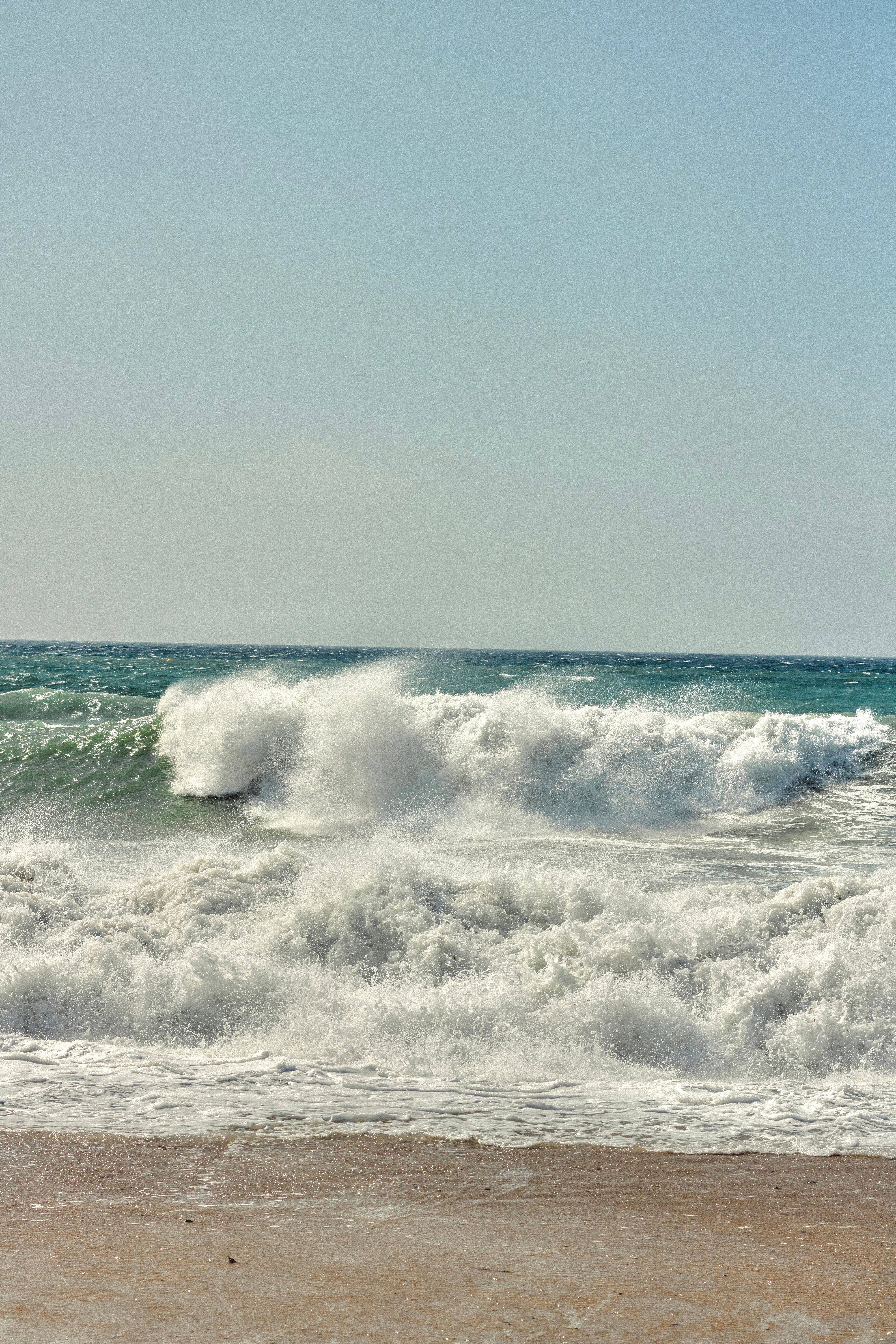 Majestic ocean waves crash onto a sandy shore under a clear sky, capturing nature's raw power.