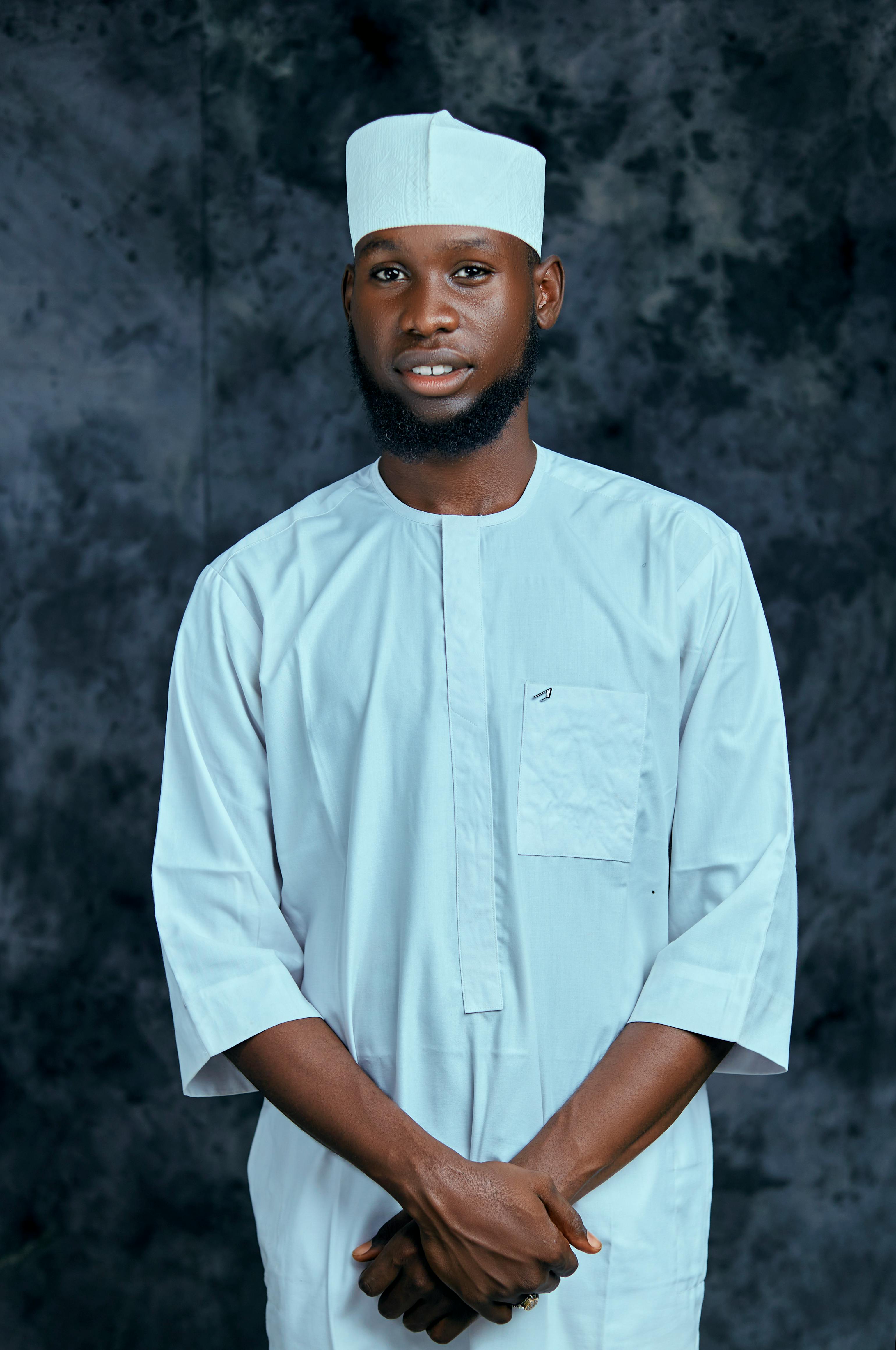 A man wearing traditional white attire with a hat against a textured background.
