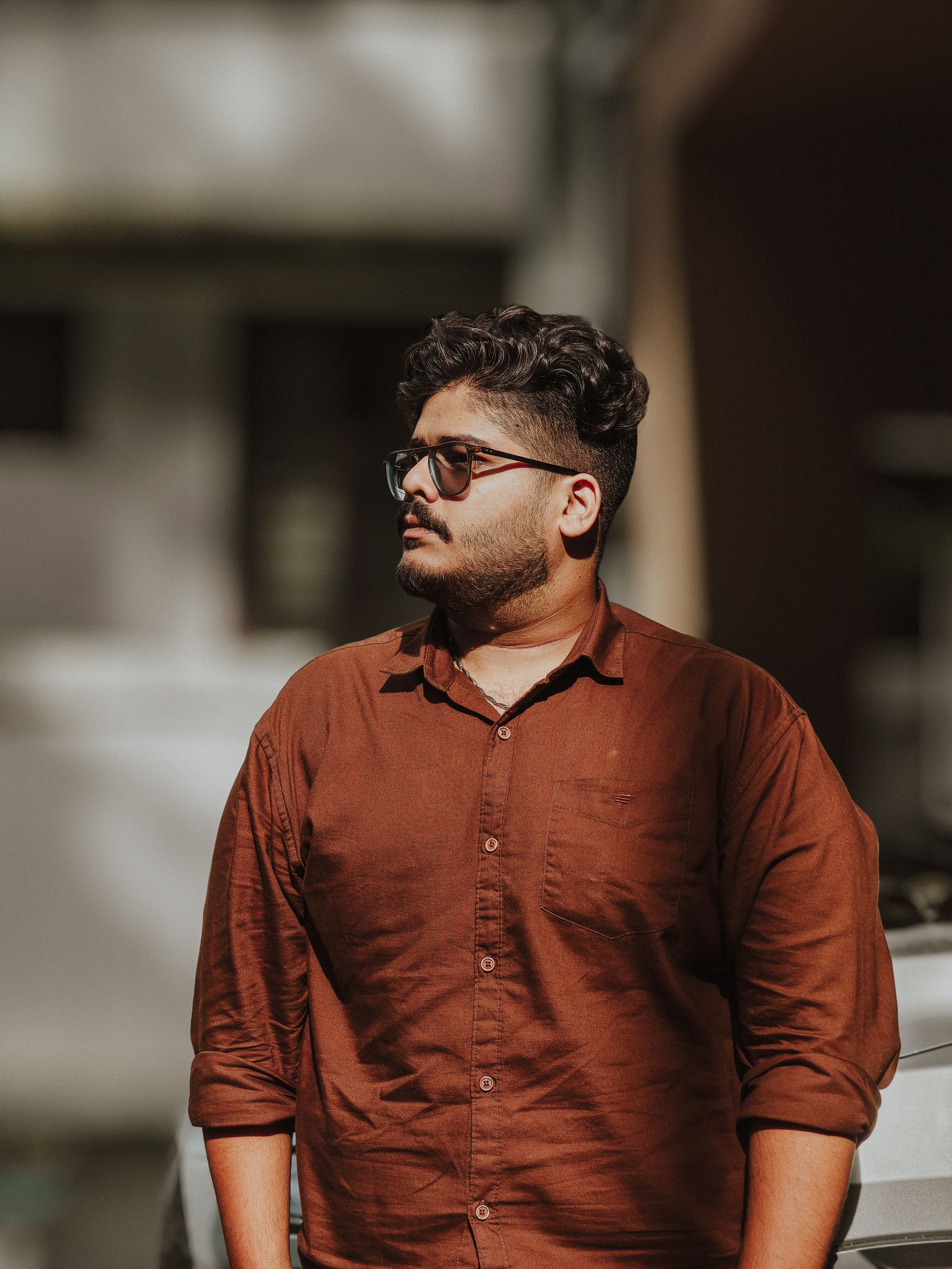 Portrait of a young man with glasses wearing a brown shirt, captured outdoors in natural light.
