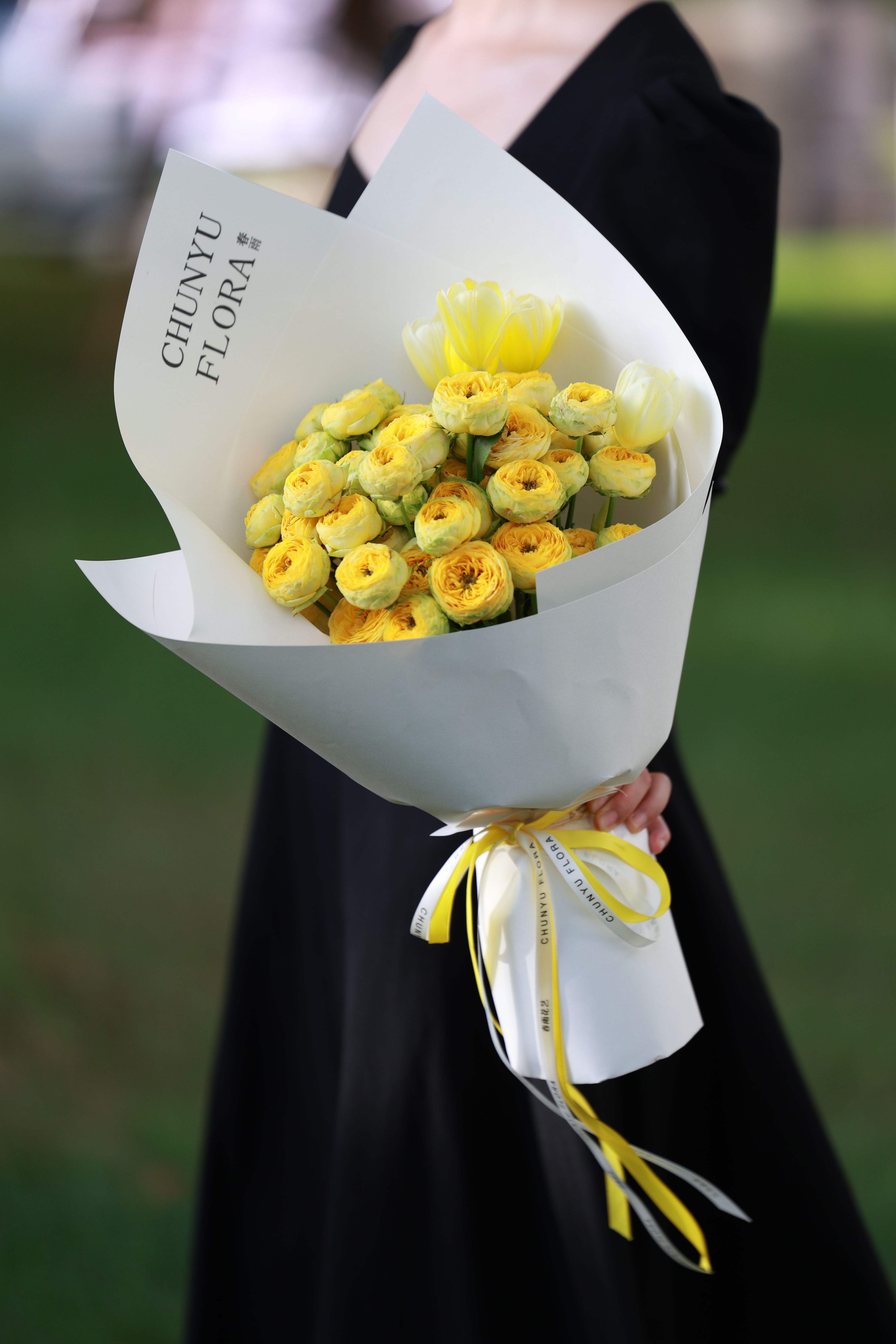 A person holding an elegant bouquet of yellow flowers wrapped in white paper outdoors.