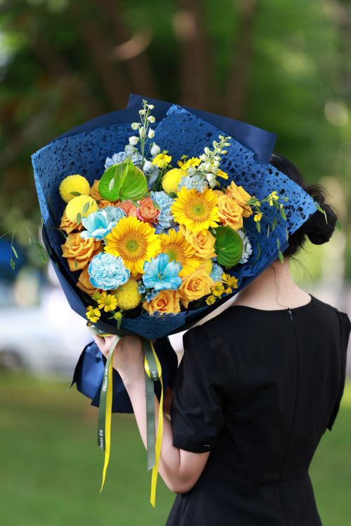 Free A woman holds a vibrant bouquet of yellow chrysanthemums and roses wrapped in blue outdoors. Stock Photo