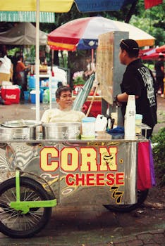 Authentic street food scene with corn cheese vendor cart under colorful umbrellas.