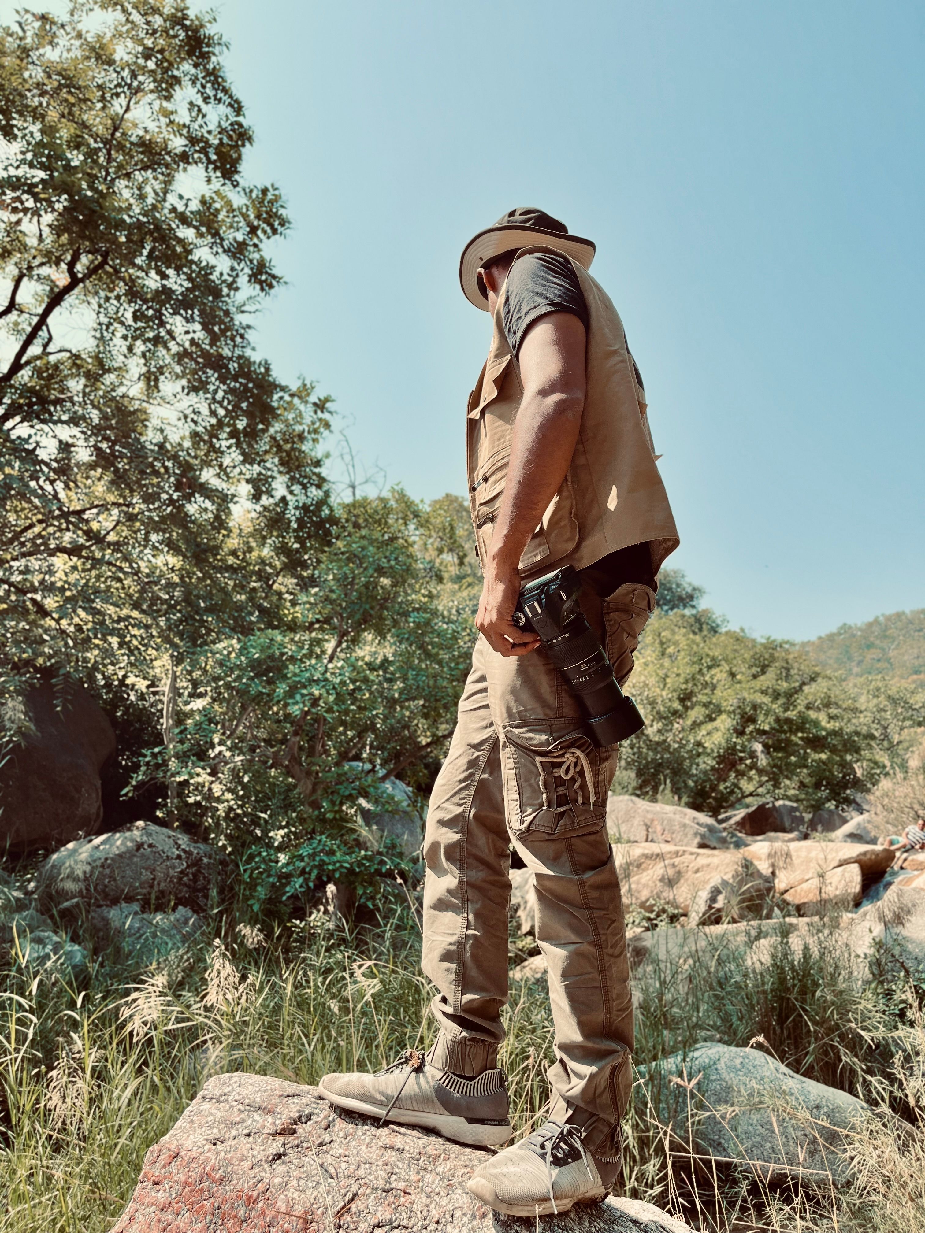 A photographer equipped for adventure, standing amidst rocky terrain in a lush outdoor setting.