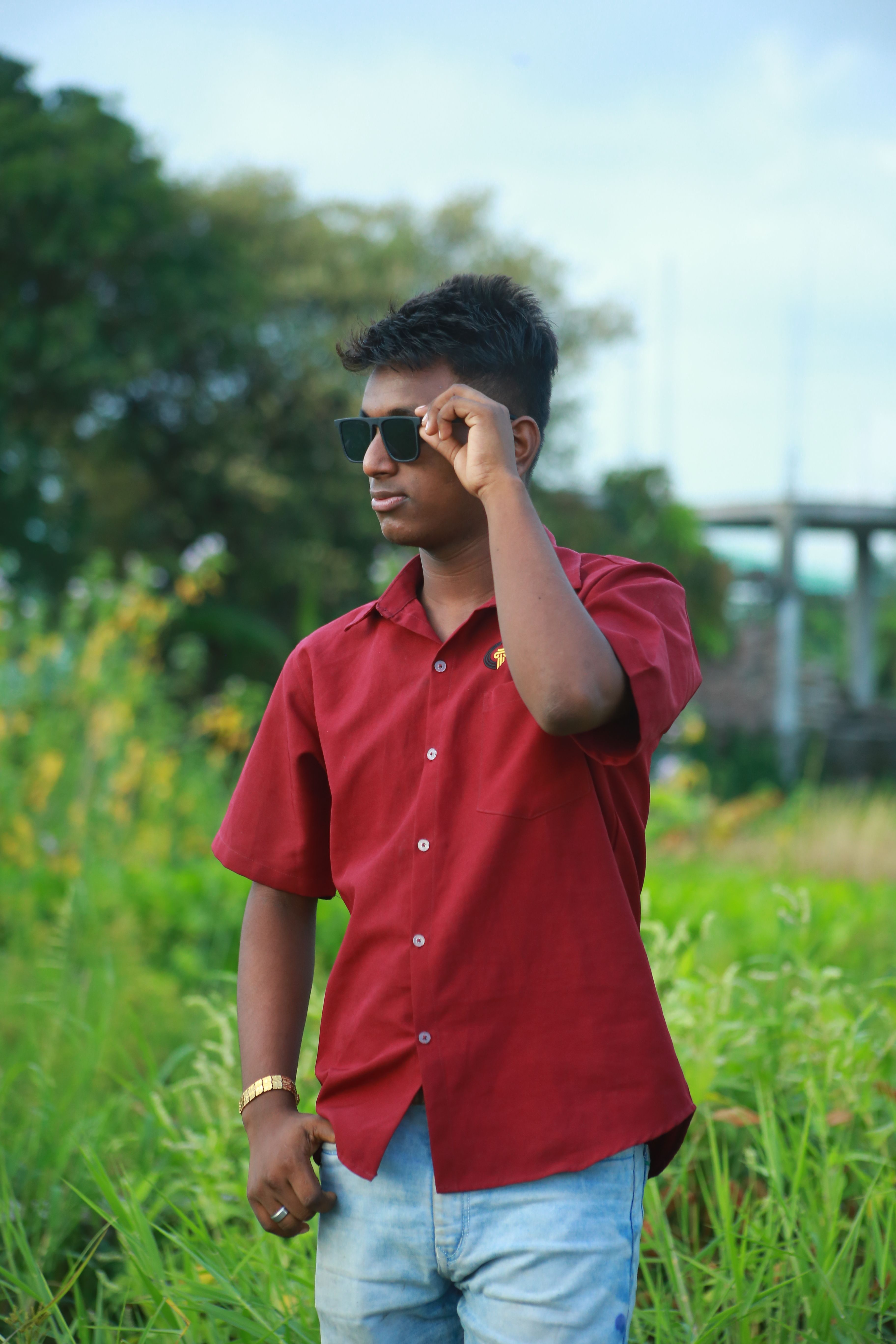 Casually dressed young man in sunglasses poses confidently in lush green outdoor setting.