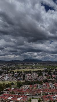 A dramatic aerial view of a Mexican city under a cloudy sky, showcasing urban landscape and distant mountains.