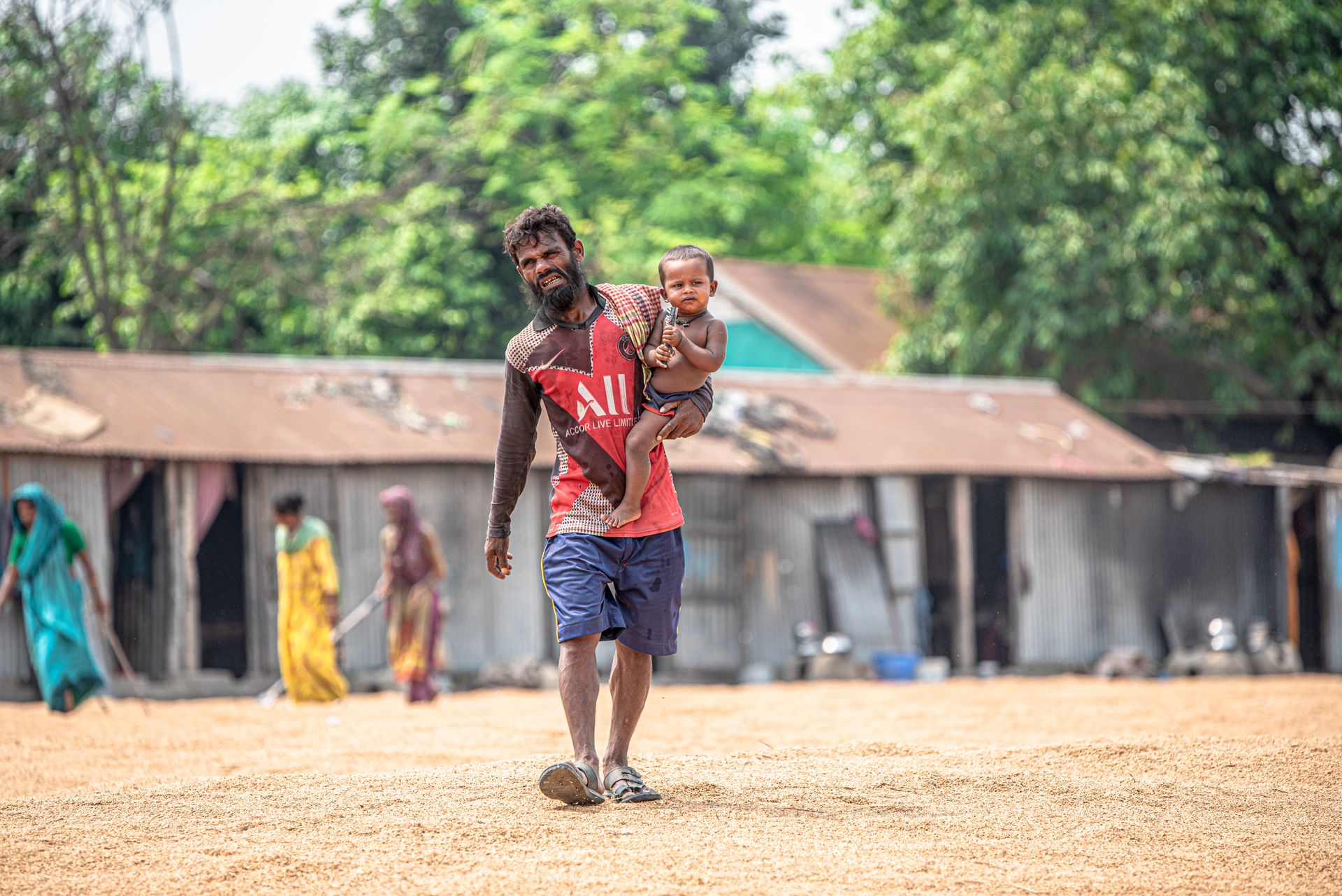 A man carries his baby through a rural village, showcasing family and daily life outdoors.