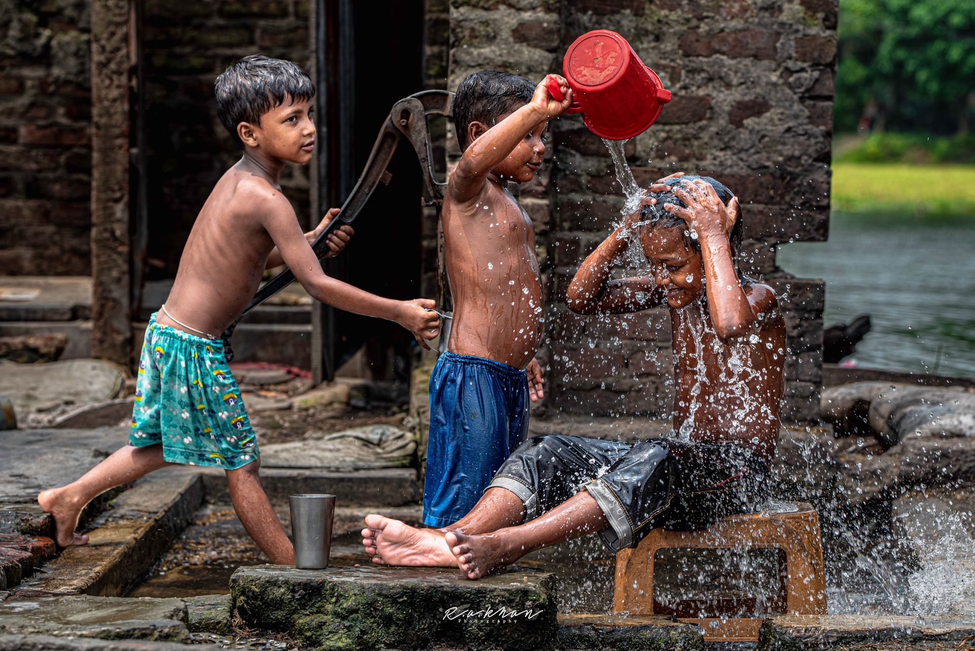 Three children playfully bathing using a hand pump outdoors beside rustic brick walls.