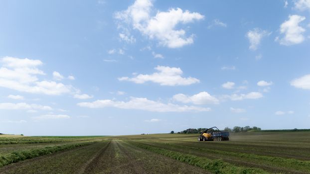 Wide-angle view of a tractor harvesting silage under a clear blue sky with scattered clouds.