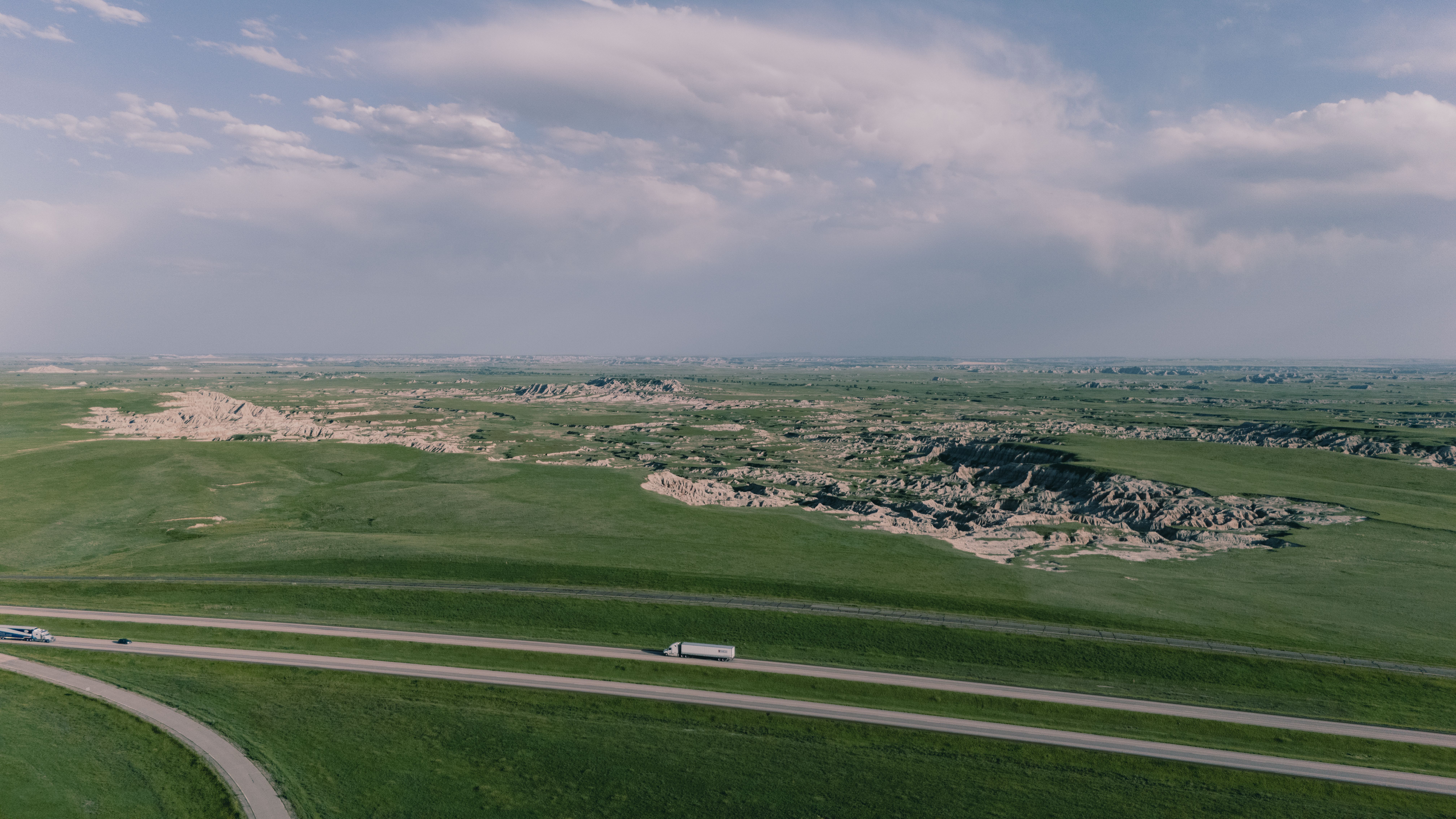Expansive aerial view of Badlands grasslands with road and distant cliffs, captured under a clear sky.
