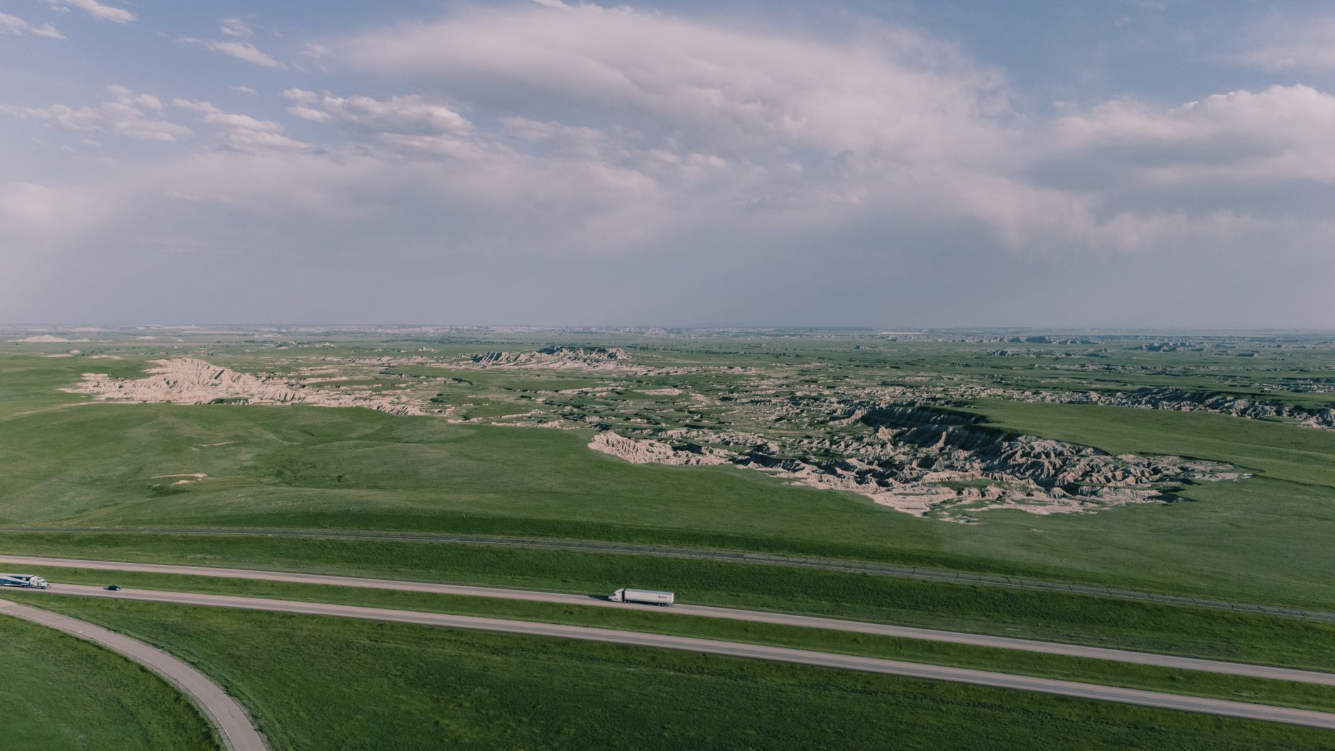 Expansive aerial view of Badlands grasslands with road and distant cliffs, captured under a clear sky.